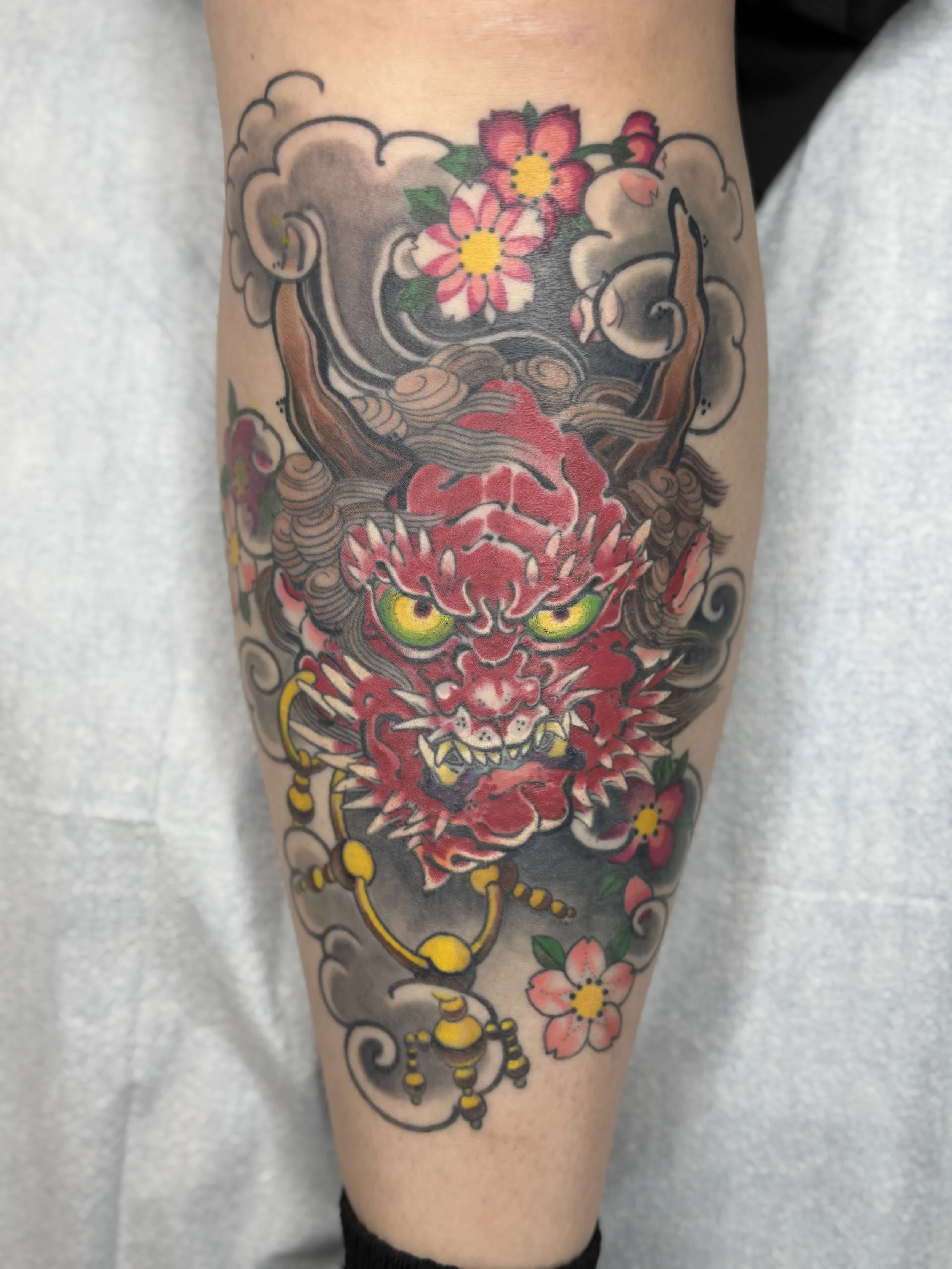 Tattoo of a stylized, fierce, red-eyed tiger with detailed fur, surrounded by pink cherry blossoms and green leaves on a person's arm.