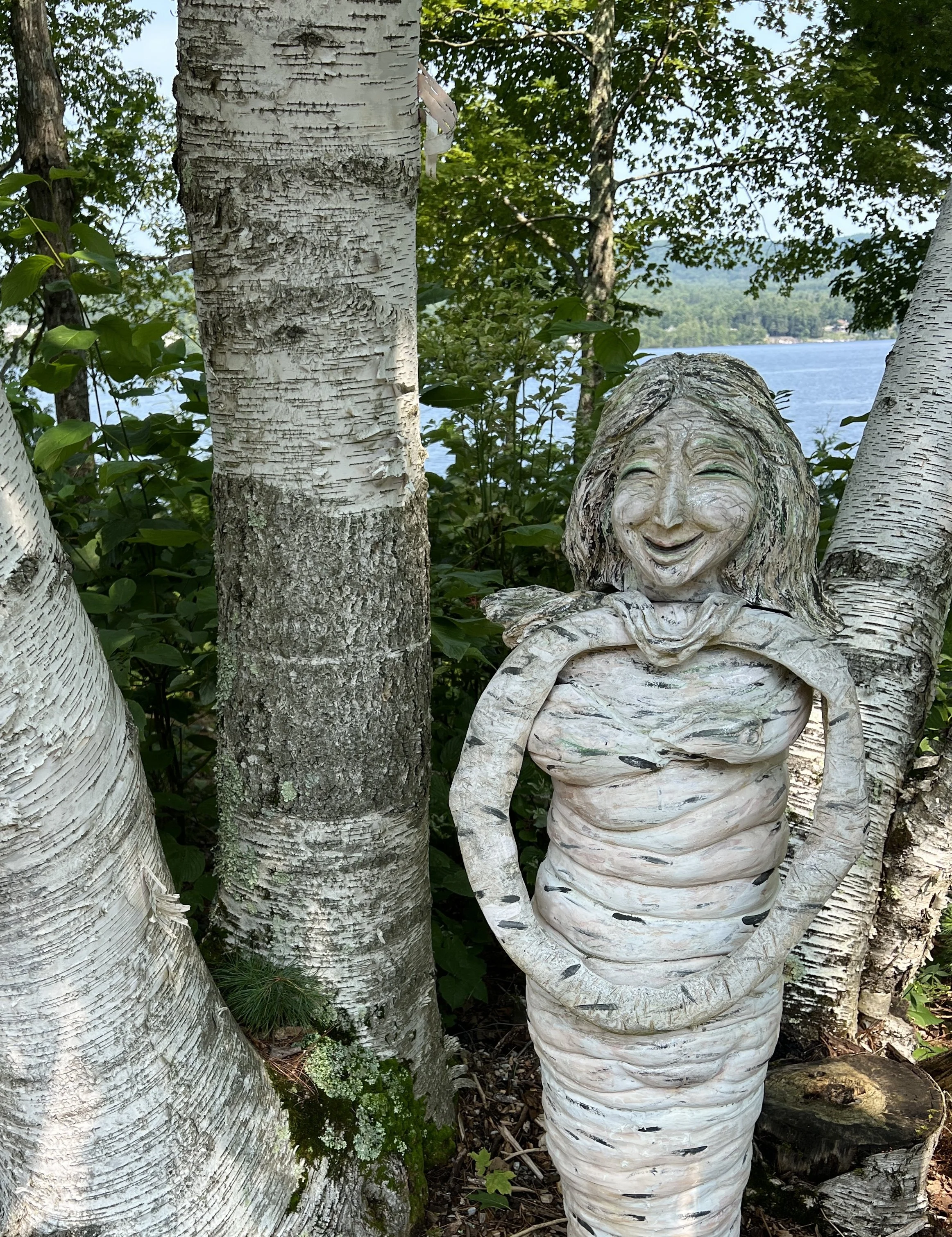 A garden scene featuring orange daylilies in the background, a wooden sculpture of a smiling woman with flowing hair and an open shirt in the foreground, and white birch trees surrounding the sculpture, near a lake.