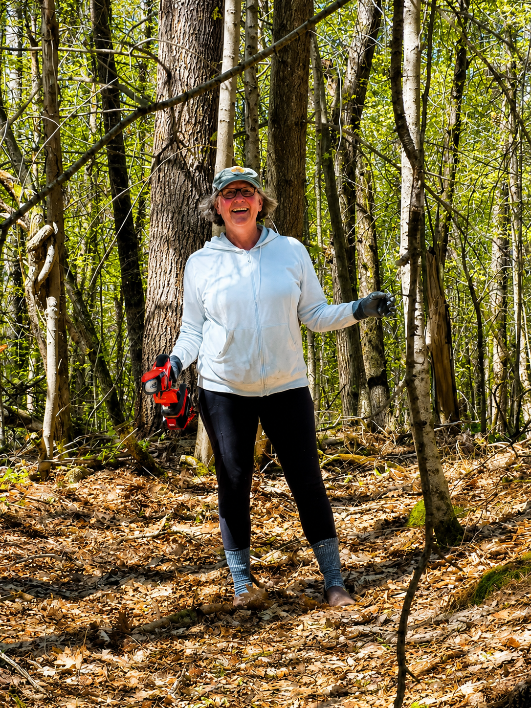A woman in a white hoodie, black leggings, and a baseball cap smiling while holding a red chainsaw in a forest surrounded by trees and fallen leaves.