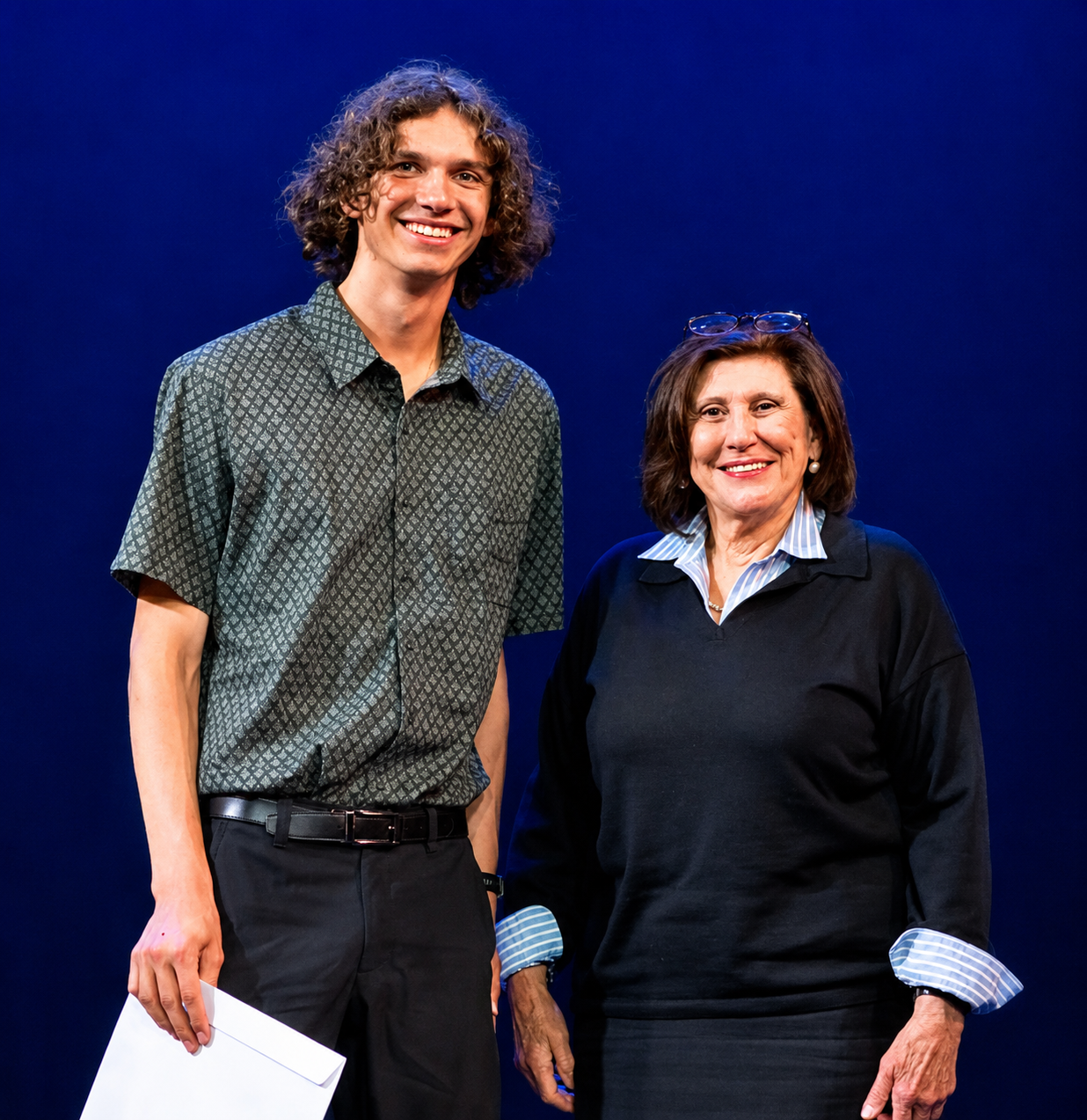 A young man with curly hair and a woman with glasses on her head, both smiling and standing against a dark blue background.