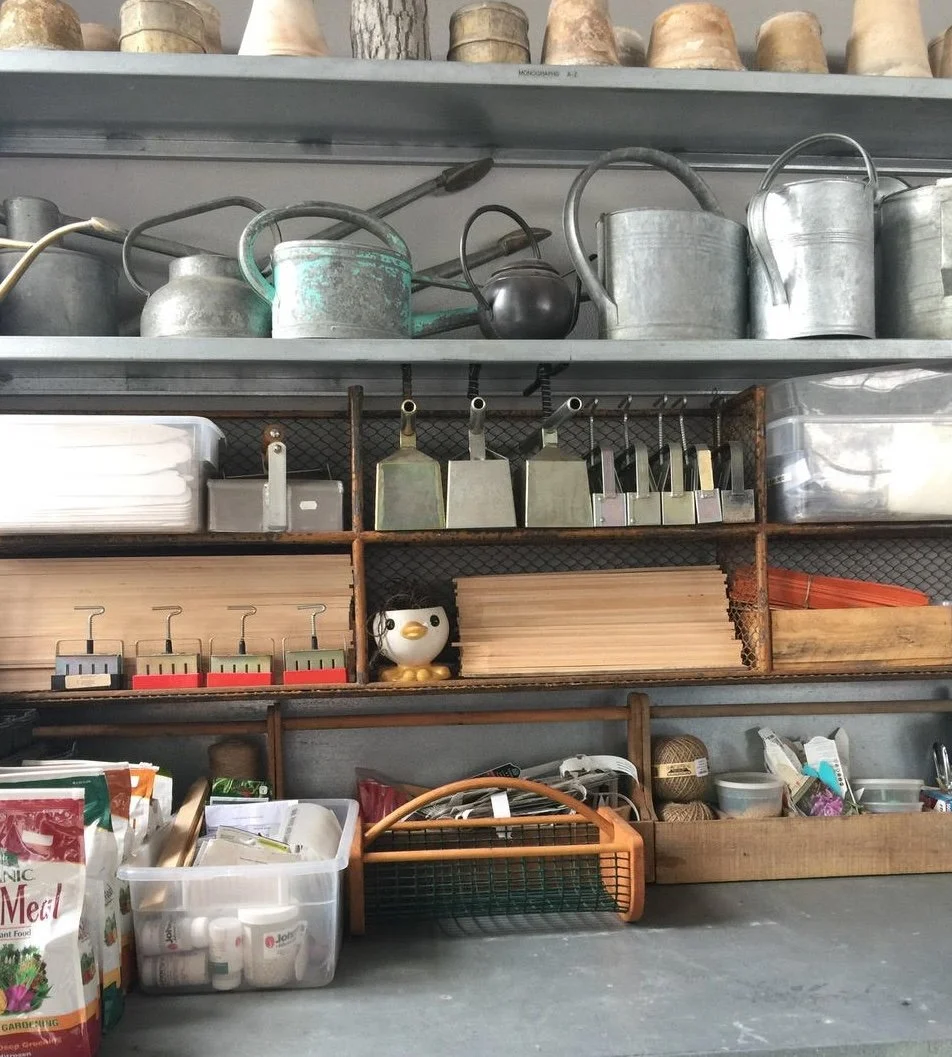 Kitchen shelf with watering cans, gardening clippers, wooden utensils, small containers, and various supplies.