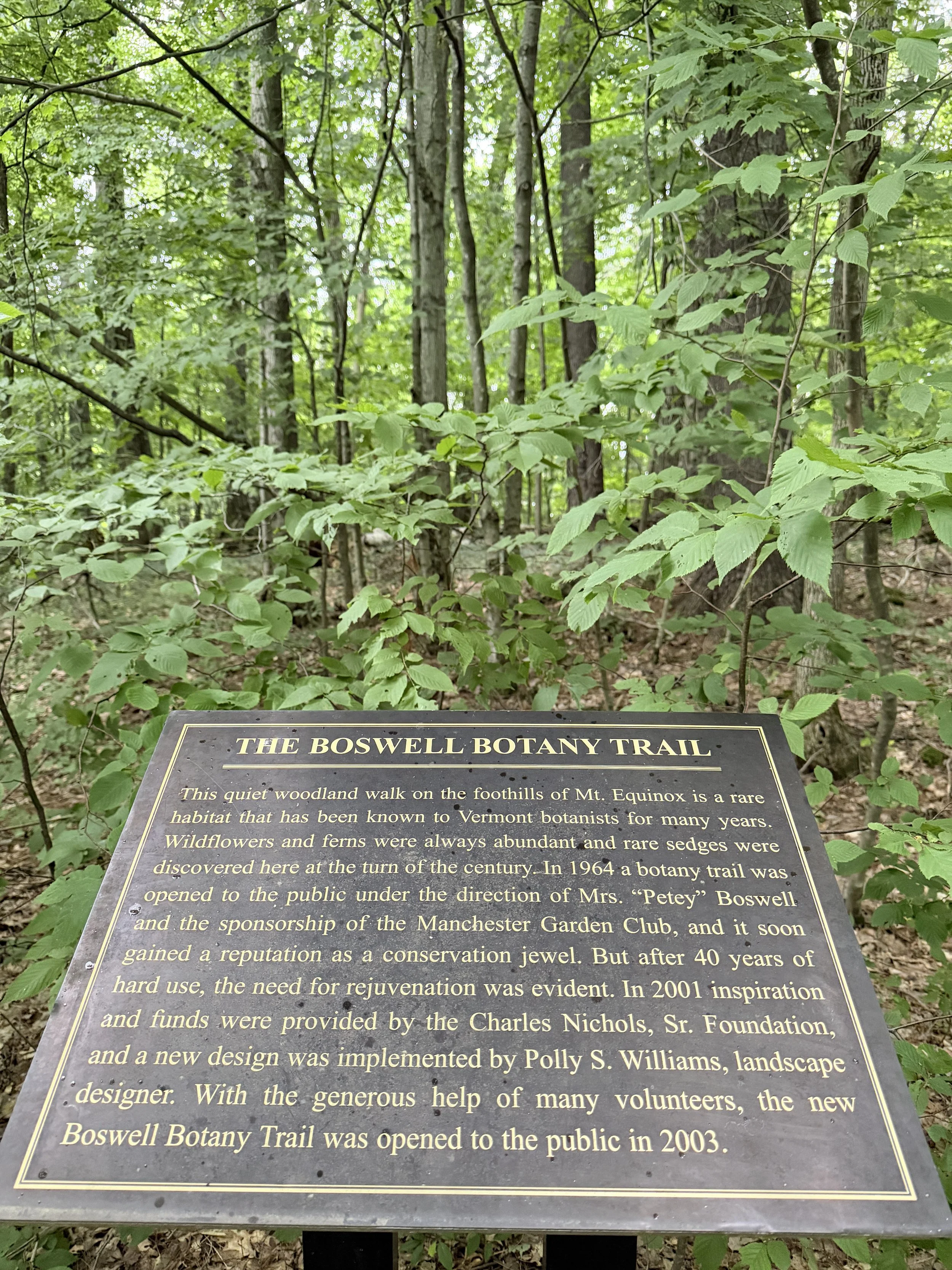 Information plaque about the Boswell Botany Trail in a wooded area, with green leaves and tree trunks in the background.
