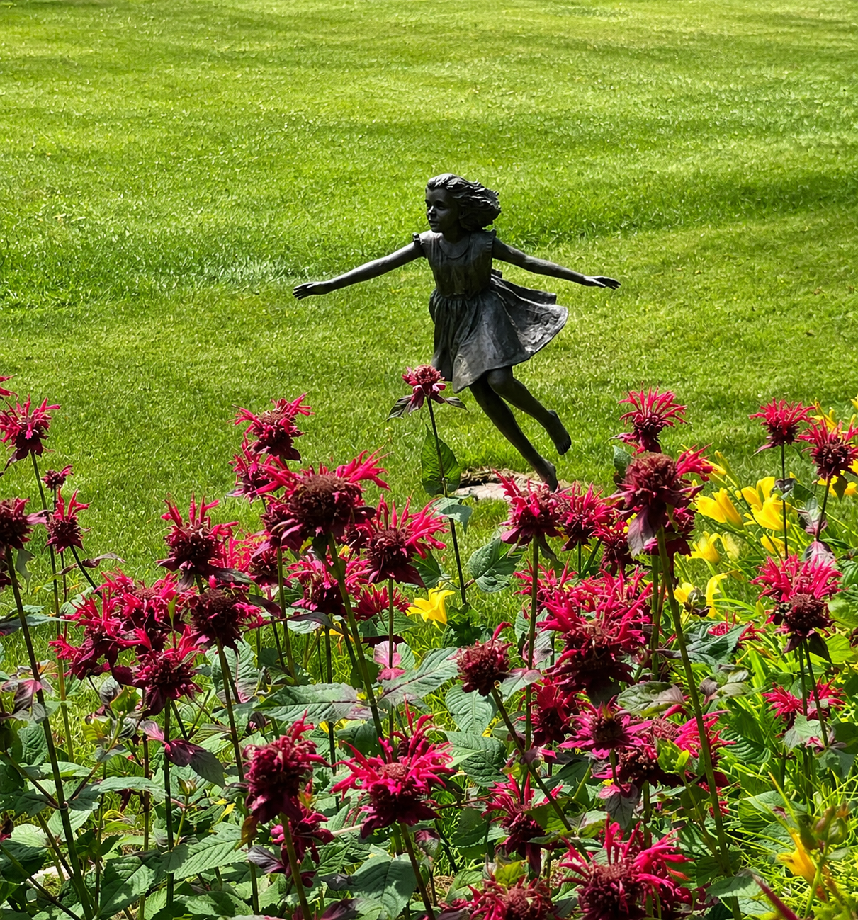Bronze sculpture of a girl in a dress, running with arms outstretched, amid pink and yellow flowers on a sunny day.