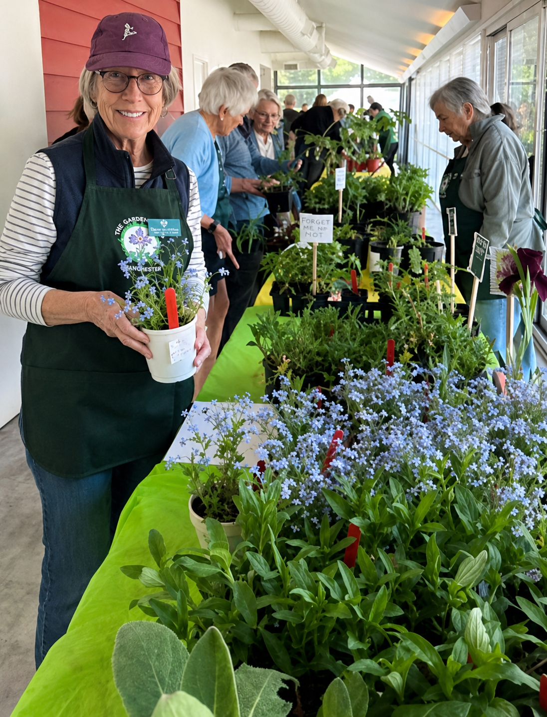 A woman wearing a purple cap, glasses, and an apron stands at a table with potted plants and flowers, smiling at the camera. Other people are browsing plants on a long table inside a bright room with large windows.