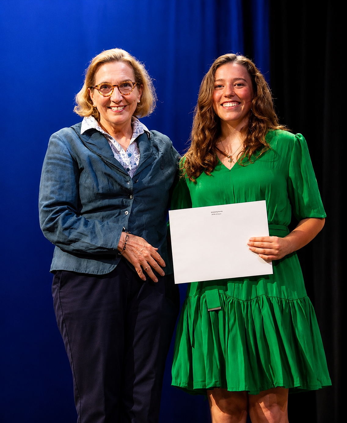 Two women standing together, one holding a certificate or diploma, smiling at an event with a dark blue curtain in the background.