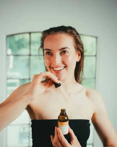 A smiling woman using a dropper to apply oil to her shoulder during a skincare routine in a bright room with large windows.