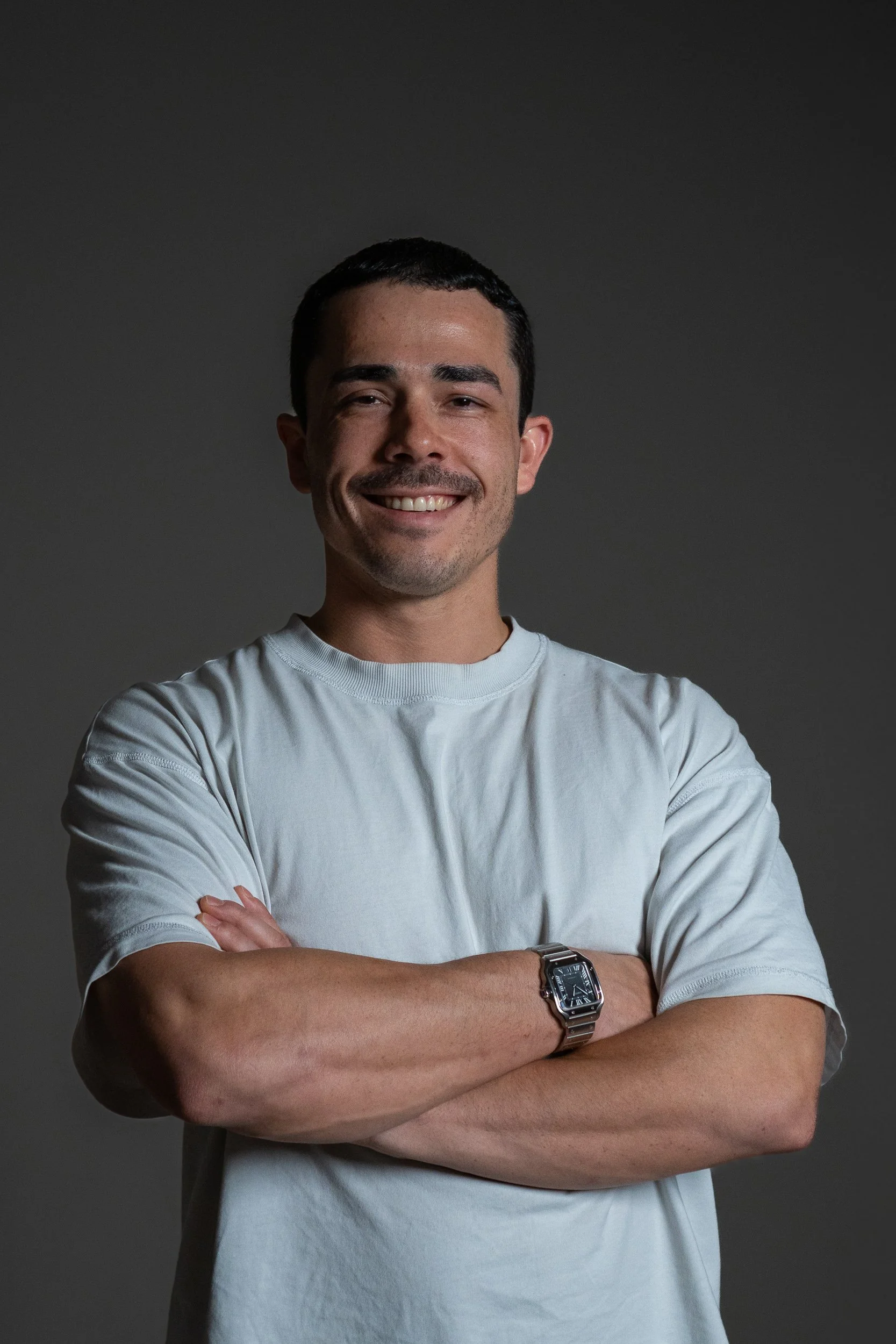Smiling man with short dark hair, crossed arms, wearing a white t-shirt and a silver watch, standing against a dark plain background.