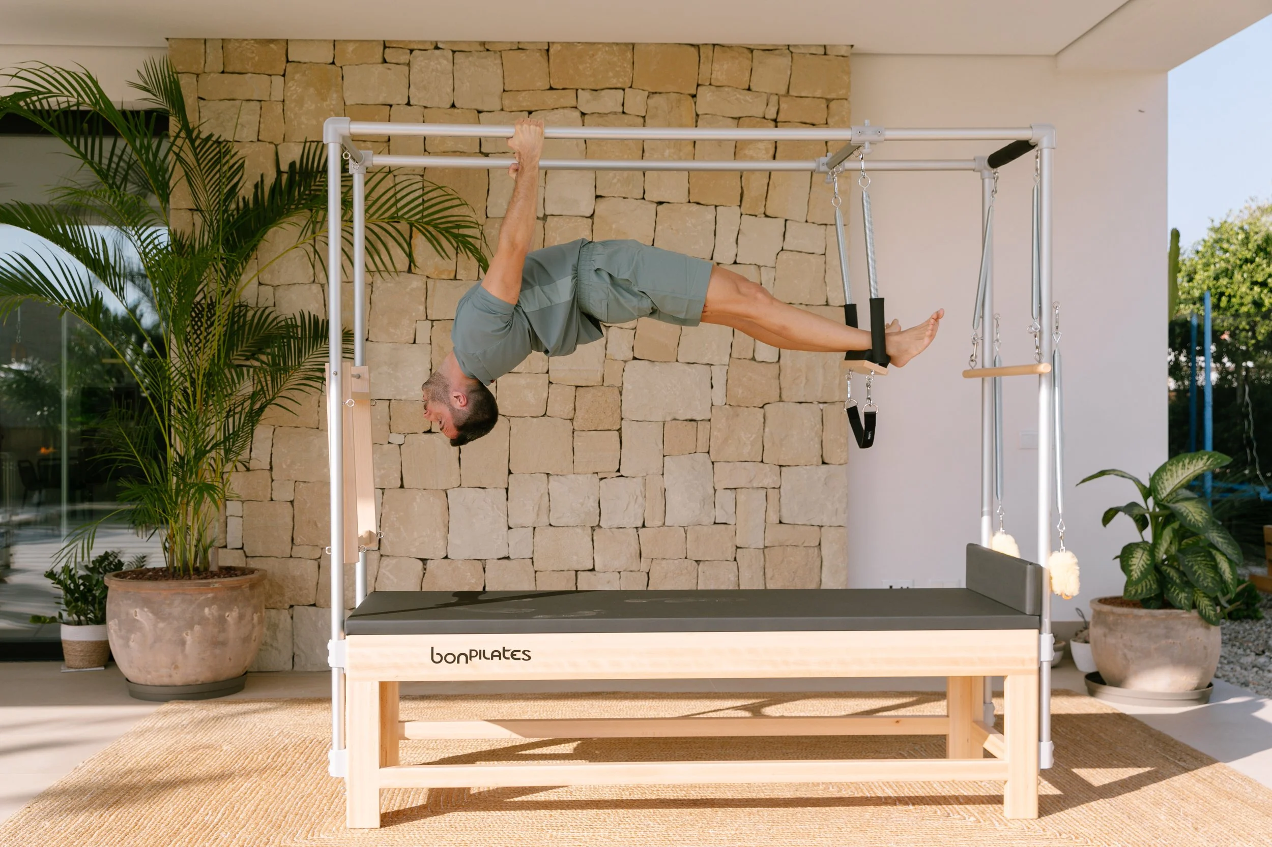 Man practicing Pilates on a reformer machine indoors, with a stone wall and green plants in the background, on a wooden platform with a beige rug.
