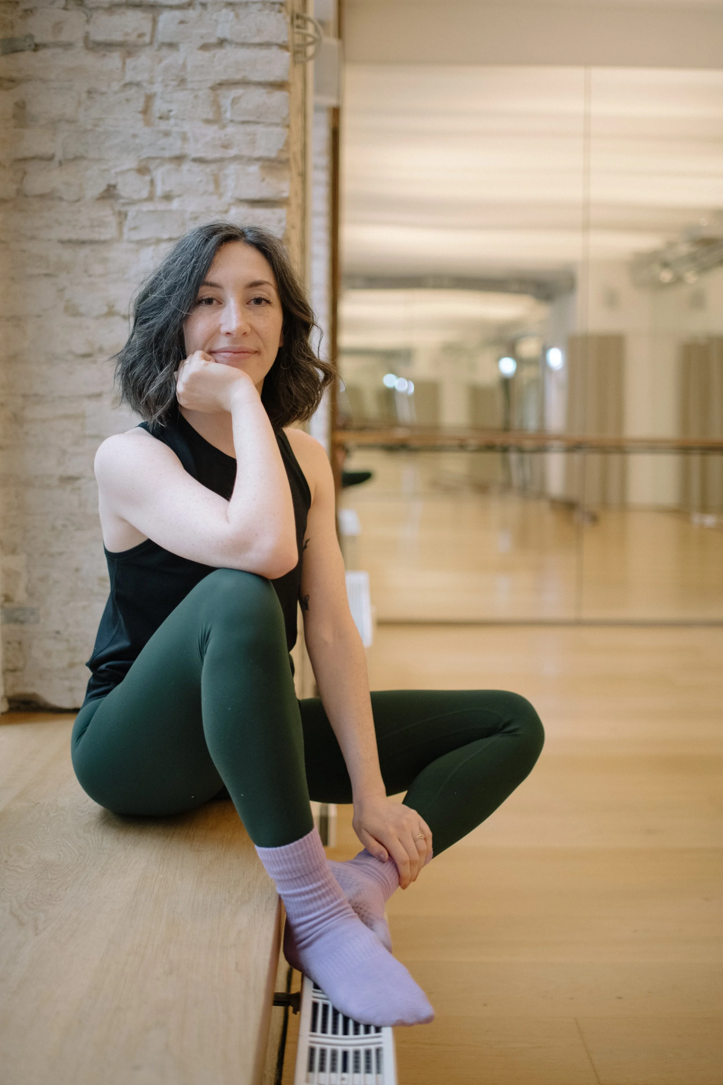A woman with dark, chin-length curly hair, wearing a black tank top and green leggings, sits on the floor of a dance studio, resting her face on her hand, with a slight smile. The studio features a mirror, ballet barre, and exposed brick wall.