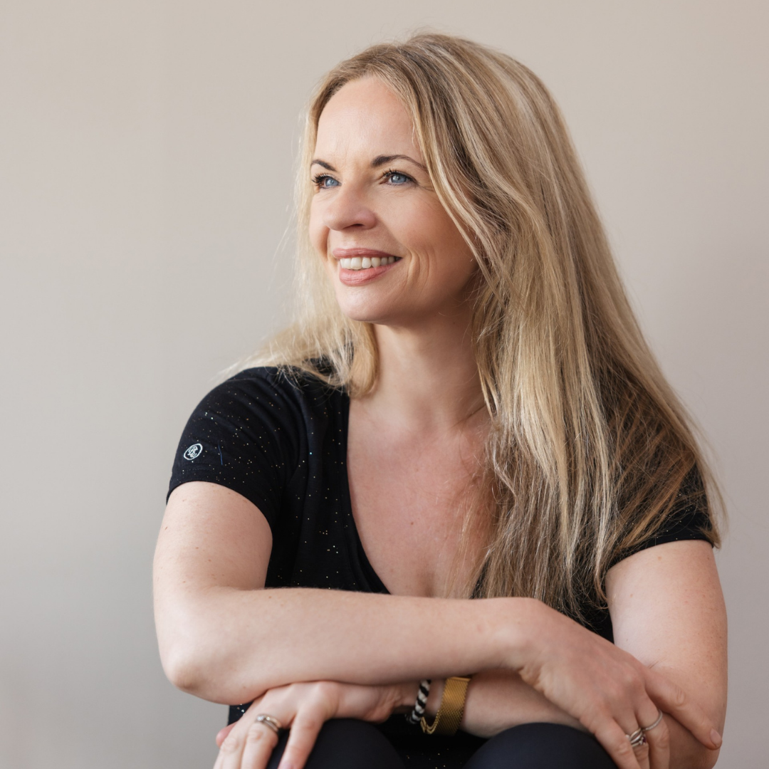 Close-up of a smiling woman with long blonde hair, wearing a black top, sitting with arms crossed, against a plain light background.