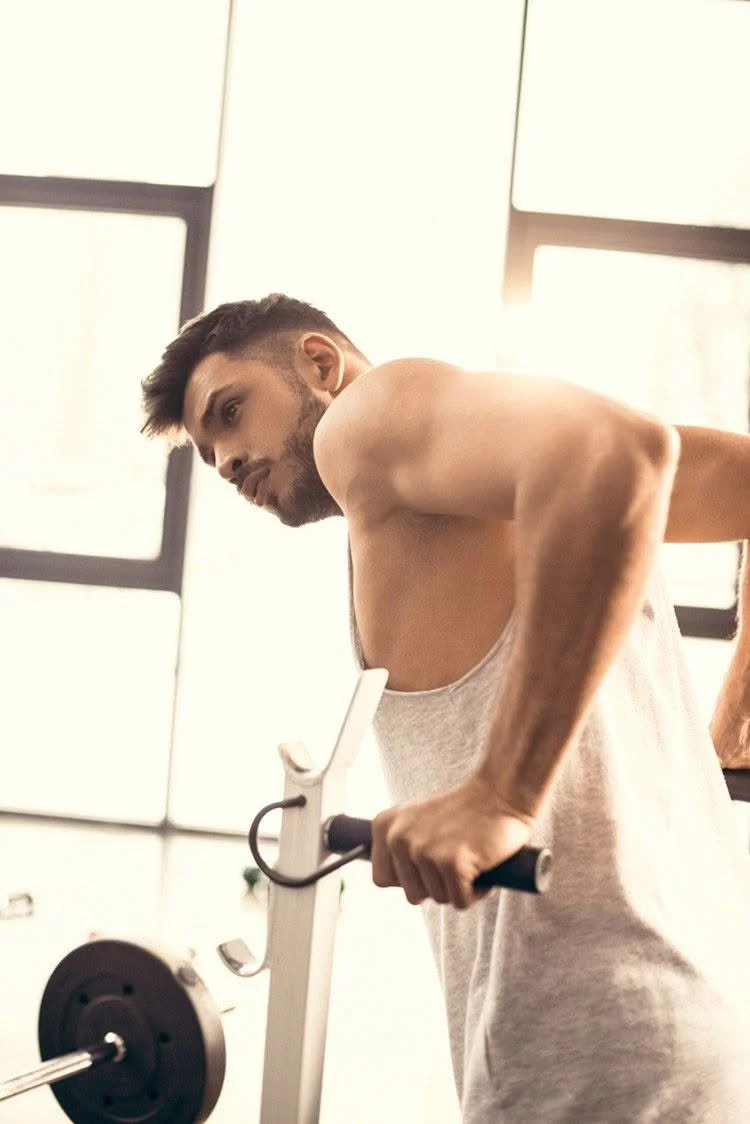 A man with a beard and short hair wearing a sleeveless shirt, working out with a rowing machine in a gym.