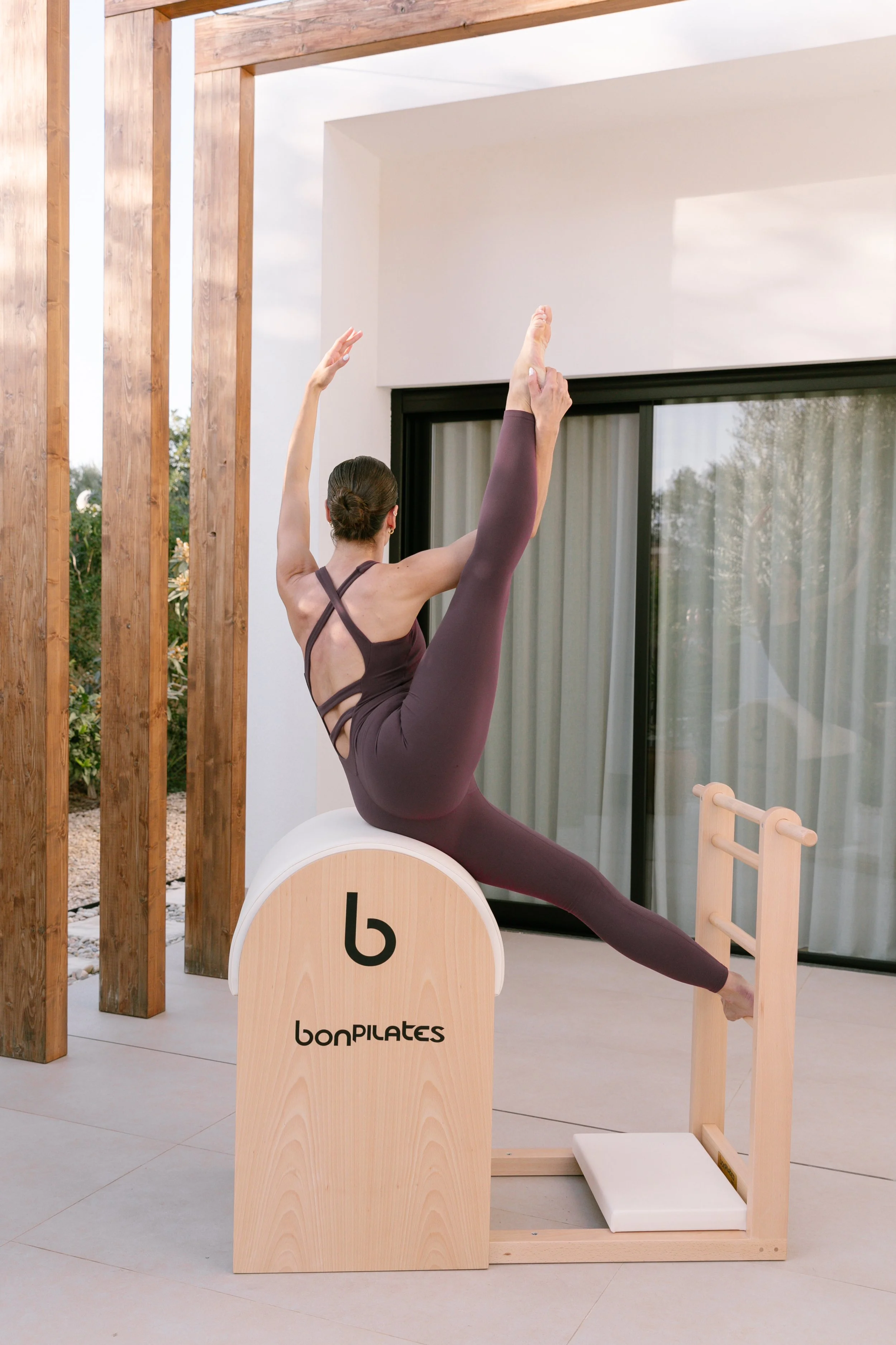 A woman performing a Pilates pose on a Pilates Ladder Barrel indoors, with large sliding glass doors and wooden decor in the background.