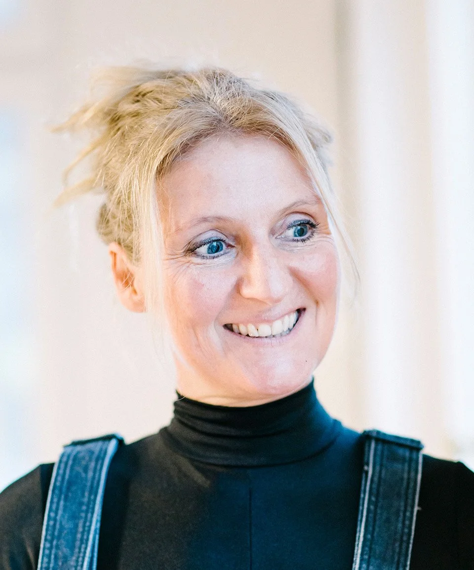 A woman with blonde hair in a loose updo, wearing a black turtleneck and denim shoulder straps, smiling and looking to her left.