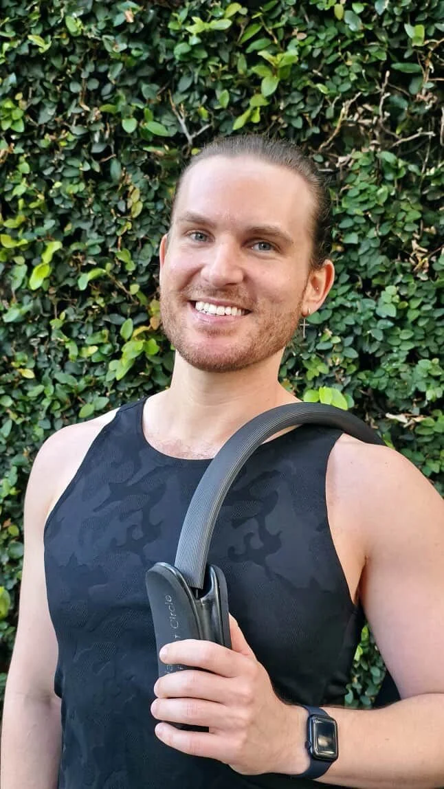 A smiling man with long hair tied back, wearing a black sleeveless athletic top, a smartwatch, and a cross earring, standing outdoors in front of a green leafy hedge, holding a black device.