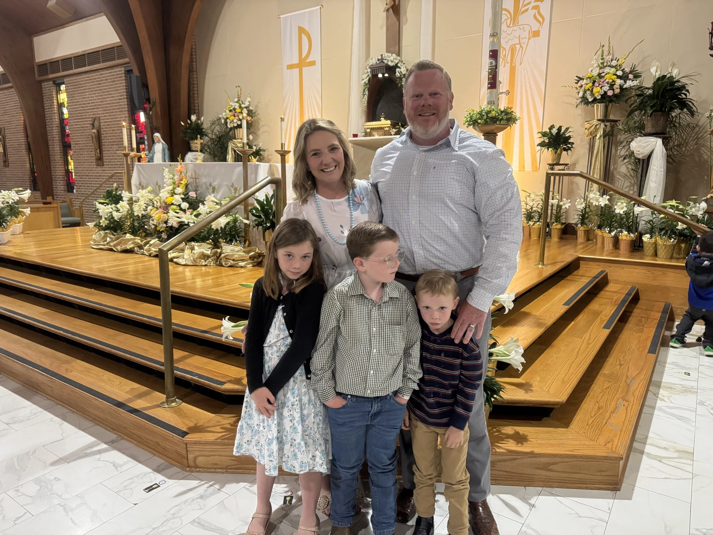 A family of six standing inside a church, smiling at the camera in front of an altar decorated with flowers and candles.