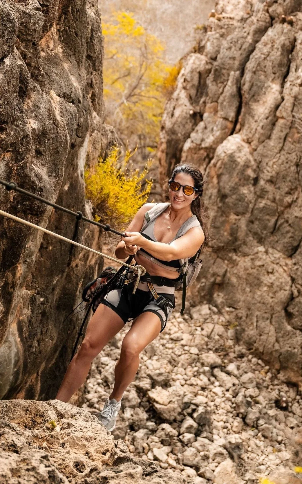 A woman rock climbing on a rugged outdoor canyon with yellow autumn trees in the background.