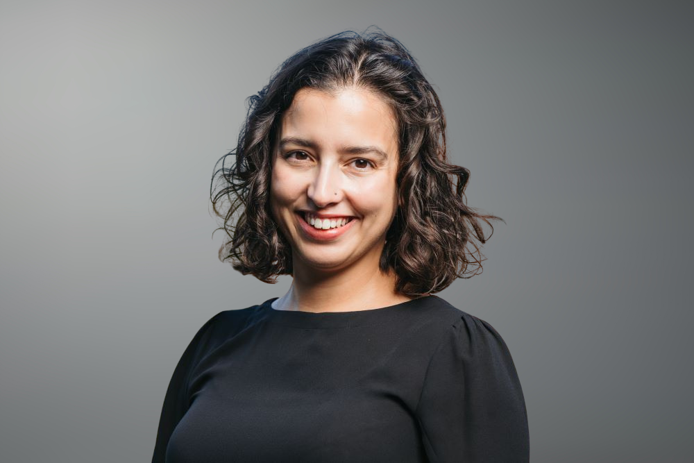 A young woman with curly brown hair, smiling, wearing a black top, standing against a plain grey background.