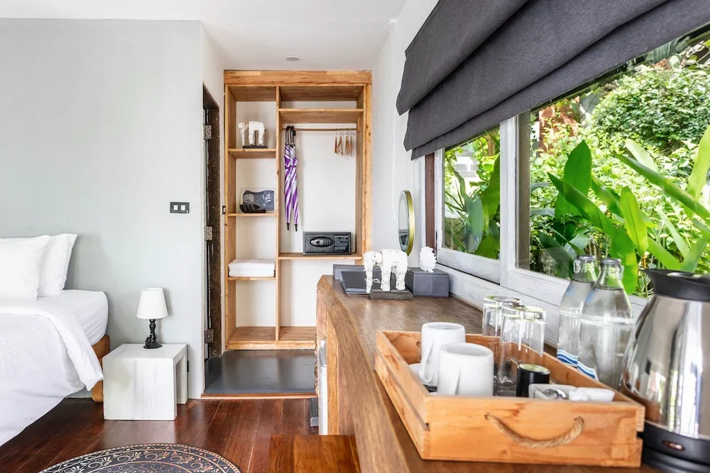 Hotel room with bed, nightstand, lamp, open closet, wooden counter with glassware, water bottles, and a window with greenery outside.