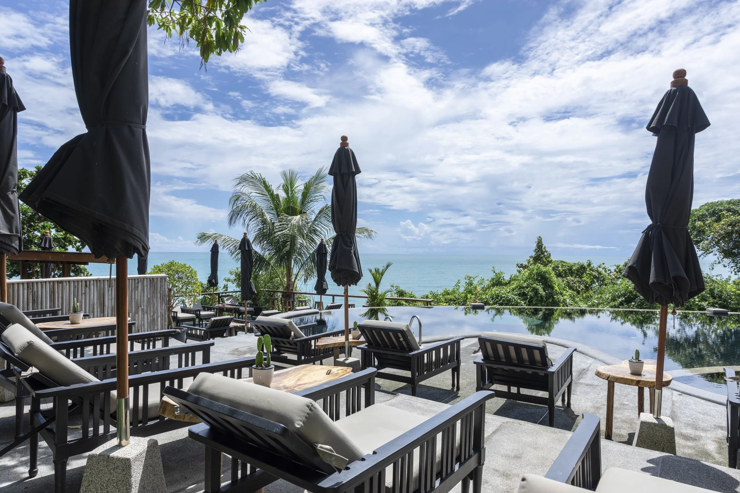 Poolside lounge chairs and tables with closed black umbrellas overlooking a tropical ocean view with blue sky and clouds.