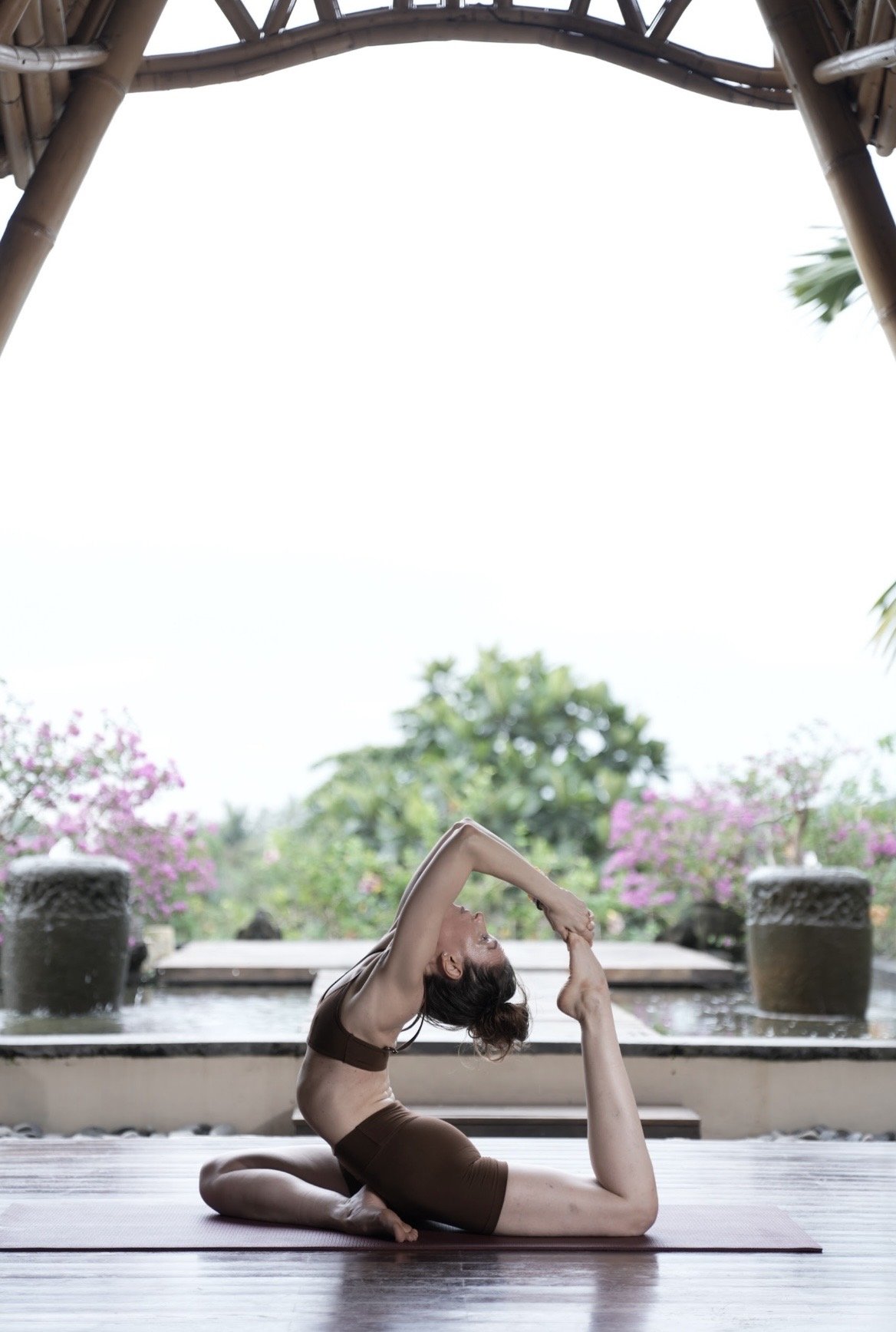 A woman practicing yoga outdoors on a mat, performing a challenging backbend pose with one arm reaching over her head and the other arm holding her foot, in a tropical garden setting with trees and pink flowering plants.