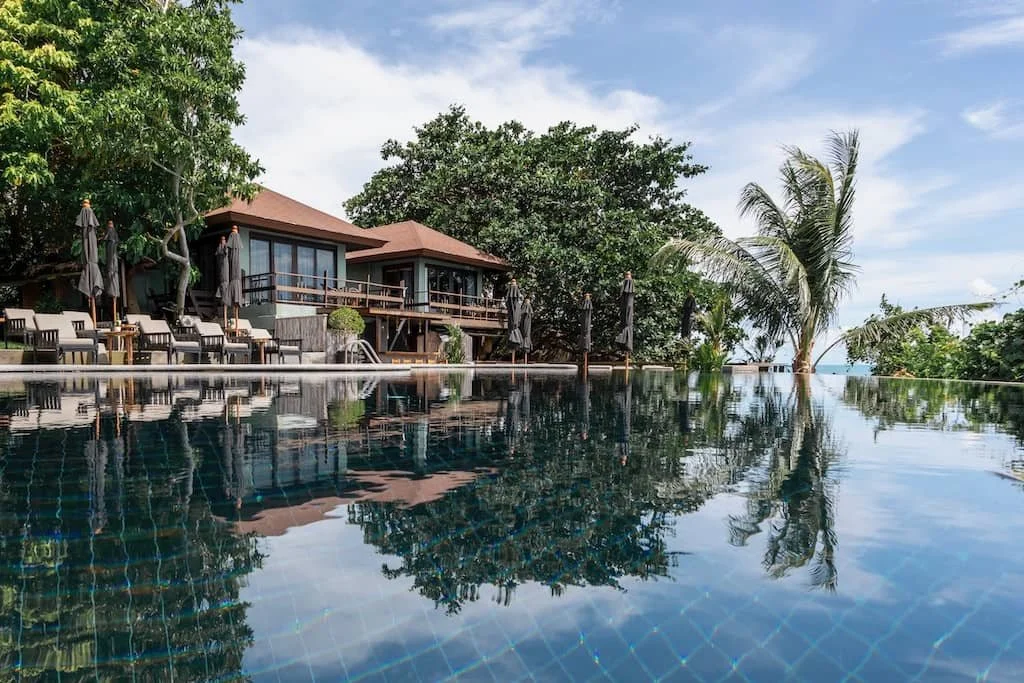 Luxury resort with an infinity pool, lounge chairs, umbrellas, and lush tropical trees near the beach.