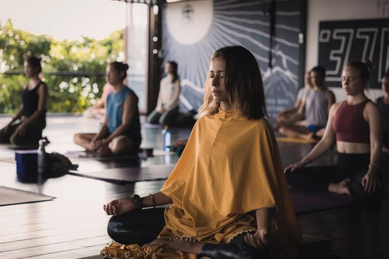 A group of women participating in a yoga or meditation class in a studio, with one woman in front wearing a yellow outfit in a meditative pose.