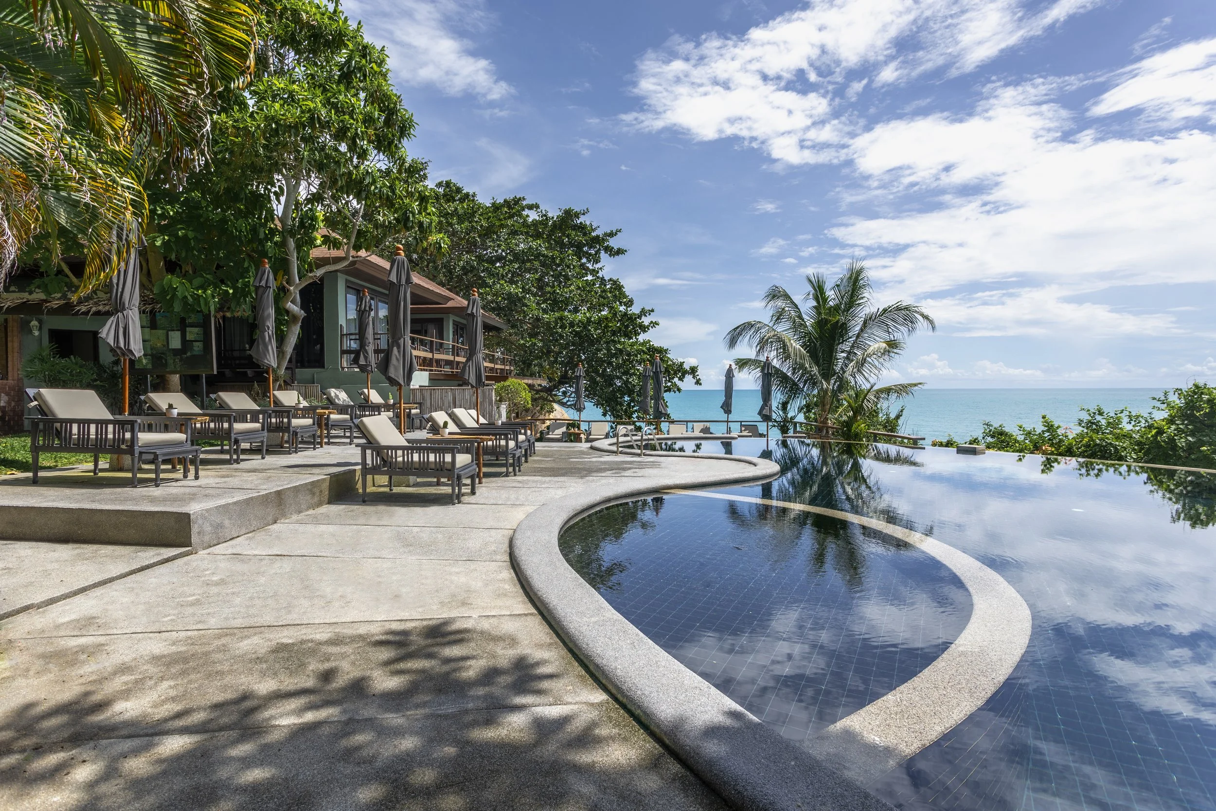 A backyard poolside area with lounge chairs, umbrellas, and a view of the ocean and blue sky with some clouds, surrounded by trees and a house.