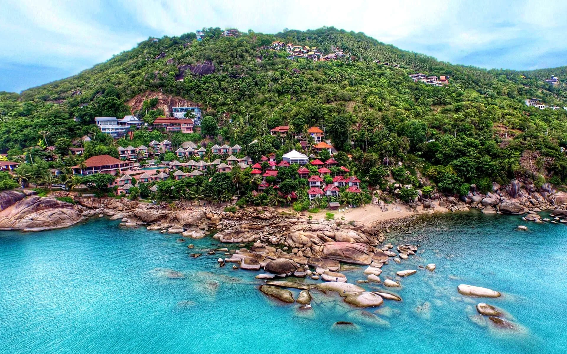 A tropical hillside scene with numerous houses and villas surrounded by green trees, overlooking a rocky coastline and turquoise ocean.