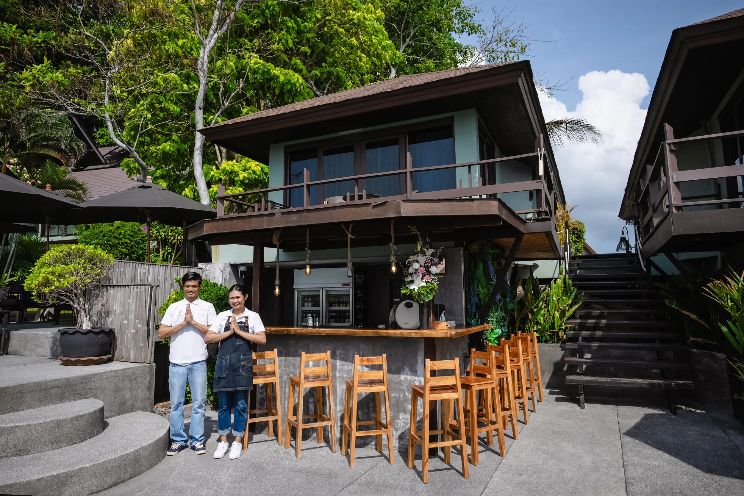 Two staff members, a man and a woman, standing outside a restaurant with wooden chairs, lush green trees, and a building with an upper balcony and staircase in the background.