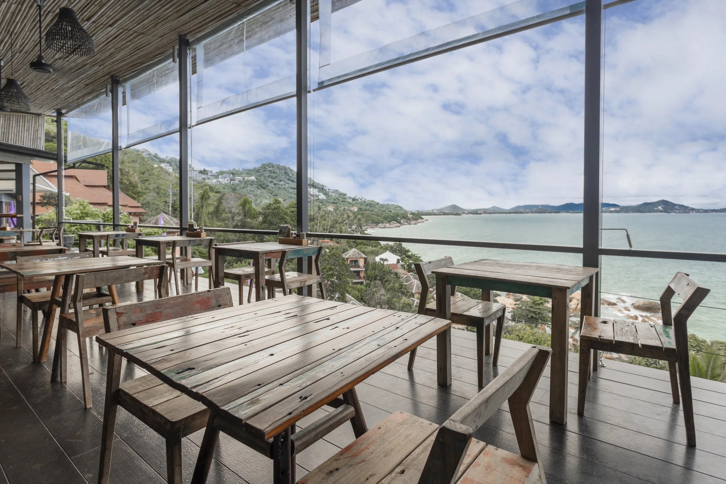 Outdoor restaurant patio with wooden tables and chairs, overlooking a beach and lush green hills, under a partially cloudy sky.