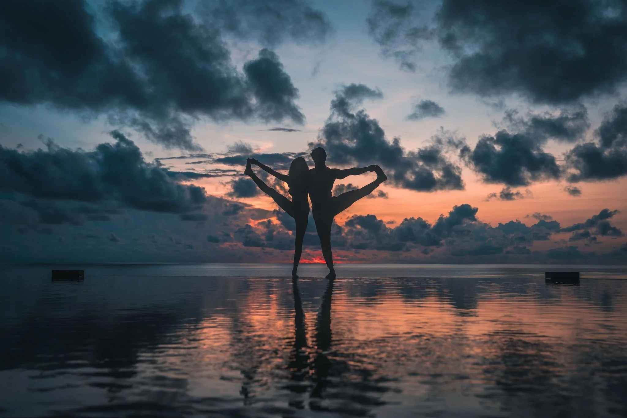 Silhouette of two people practicing partner yoga on the beach at sunset, with clouds in the sky and water reflecting the scene.