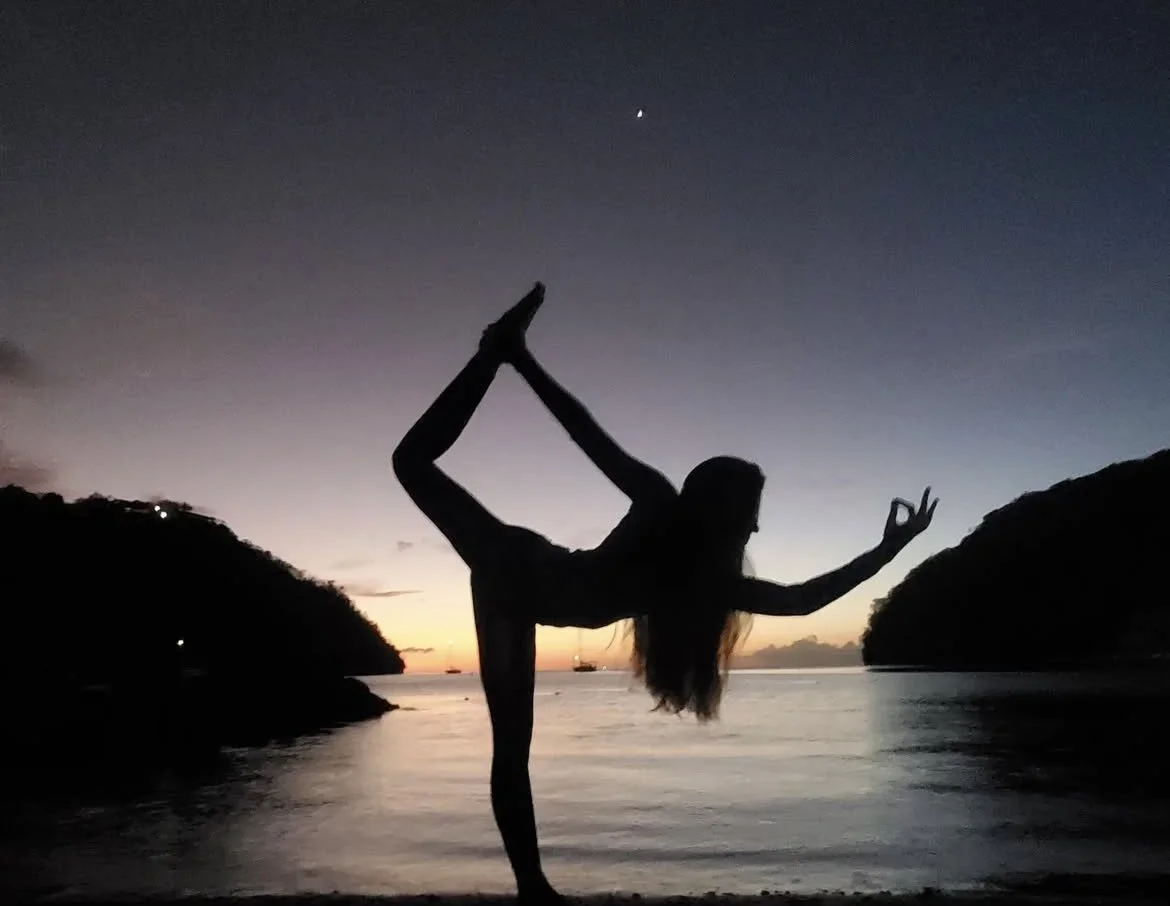Silhouette of a woman performing a yoga pose on a beach at sunset, with the sky transitioning from orange to dark blue and distant islands visible.