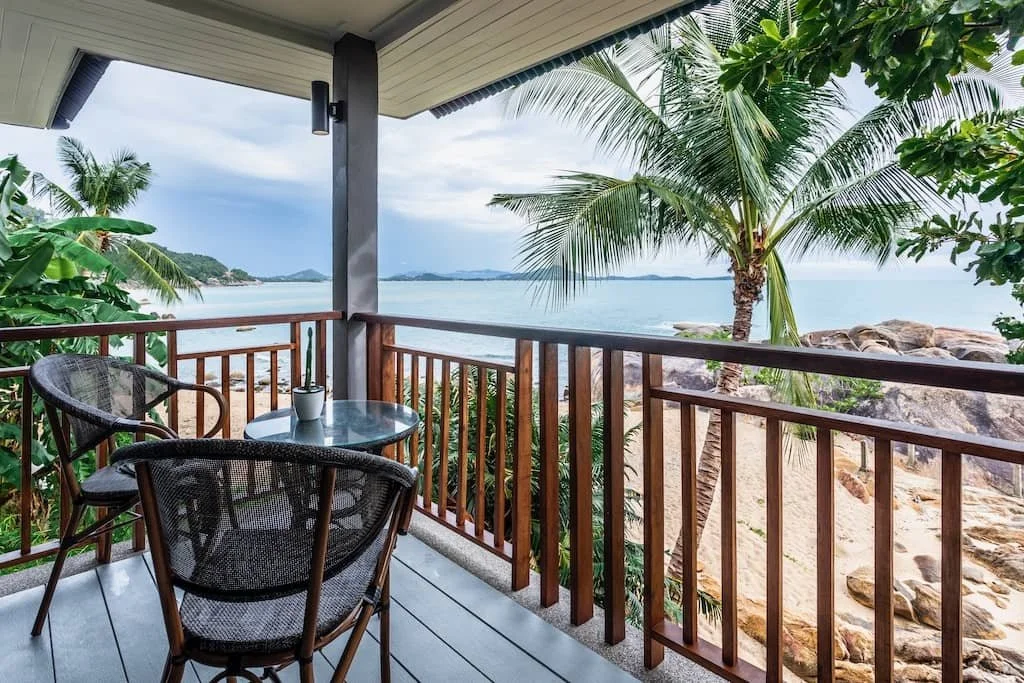 Balcony overlooking tropical beach with palm trees, rocks, and water, with a small table and two chairs on the porch.