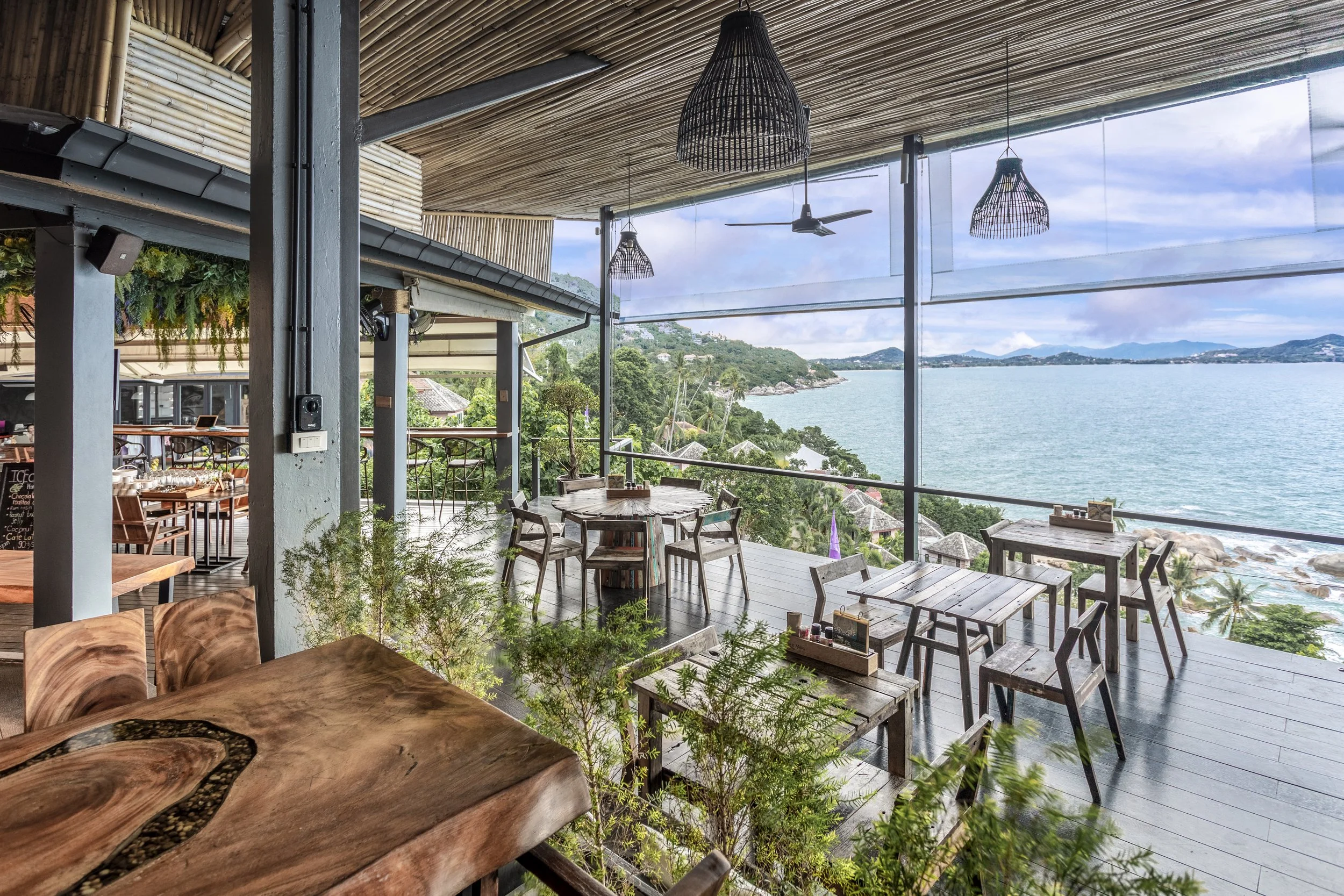 An open-air restaurant or lounge with wooden tables and chairs, overlooking a lake with mountains in the distance, under a bamboo ceiling with hanging woven light fixtures.