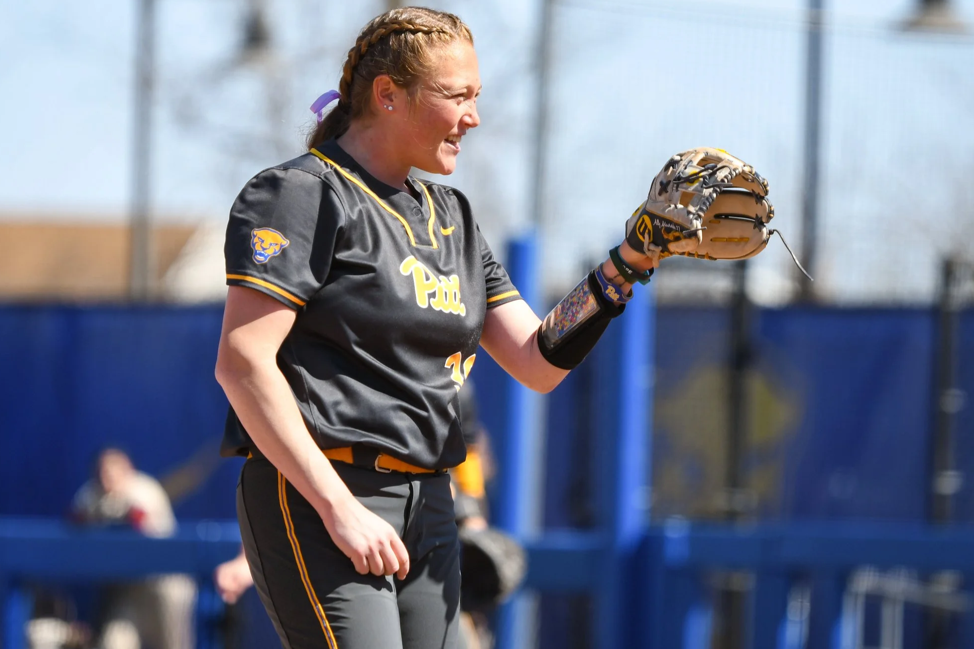 A female softball player with braided hair wearing a black and yellow Pitt Panthers uniform on the field, holding a glove and smiling.