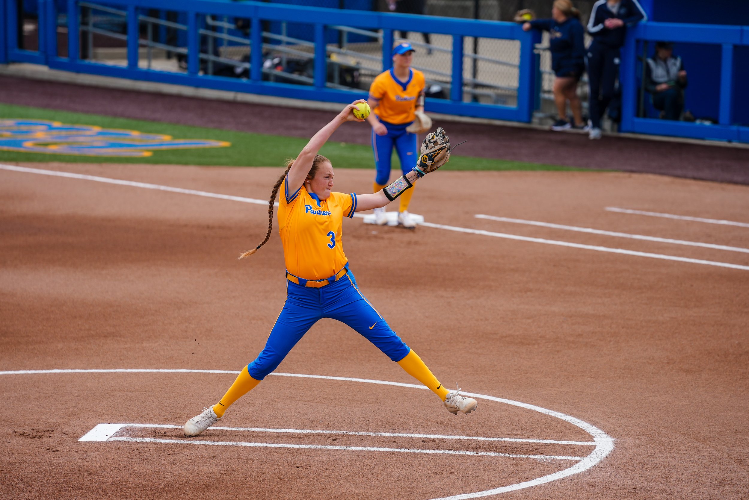 A softball player in a yellow uniform and blue pants is about to pitch the ball during a game, with her body stretched forward and her arm extended.