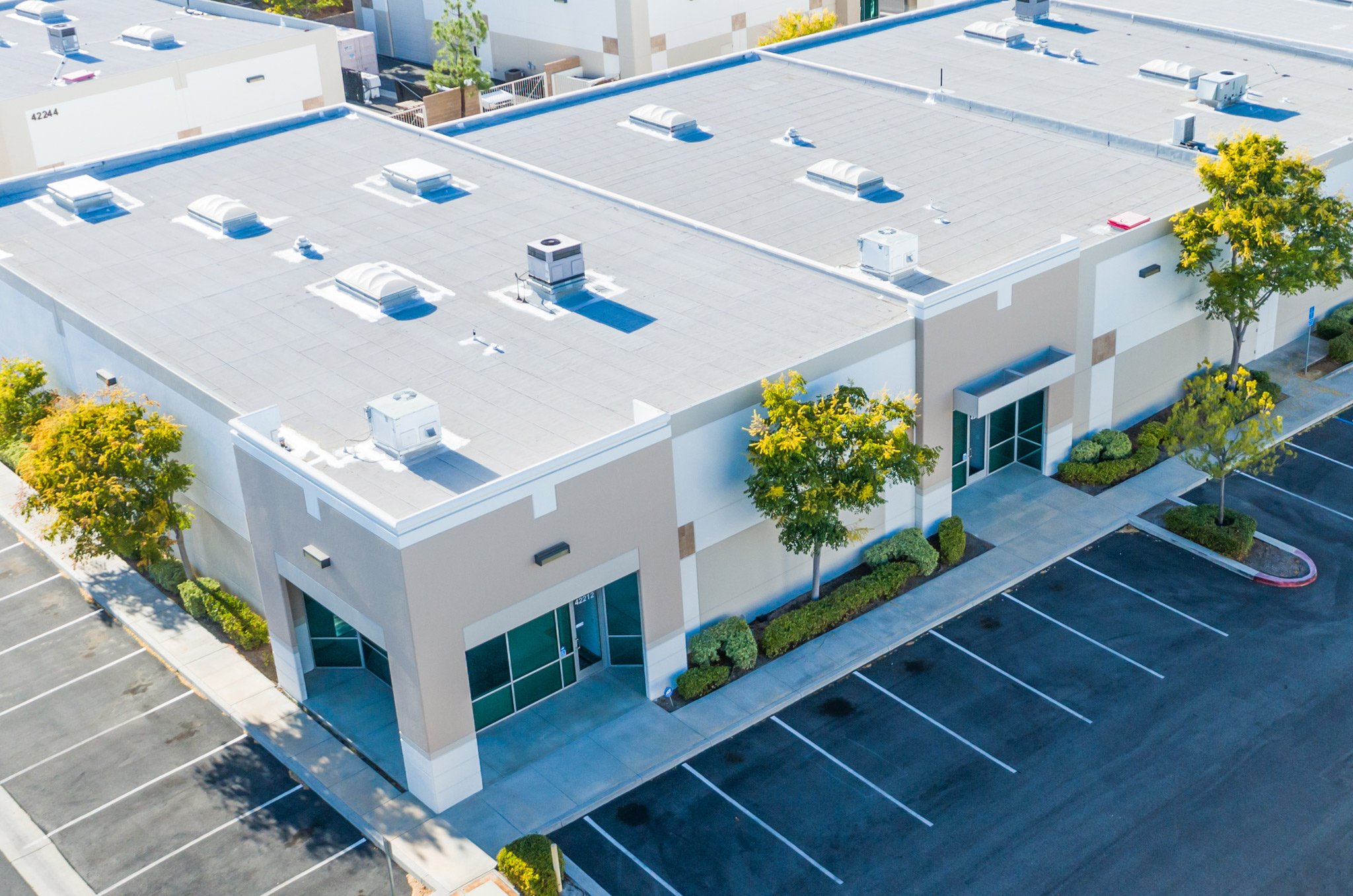 An aerial view of a commercial building with a flat roof, featuring multiple vents and HVAC units. The building has large glass windows and is surrounded by trees, with a parking lot in front.