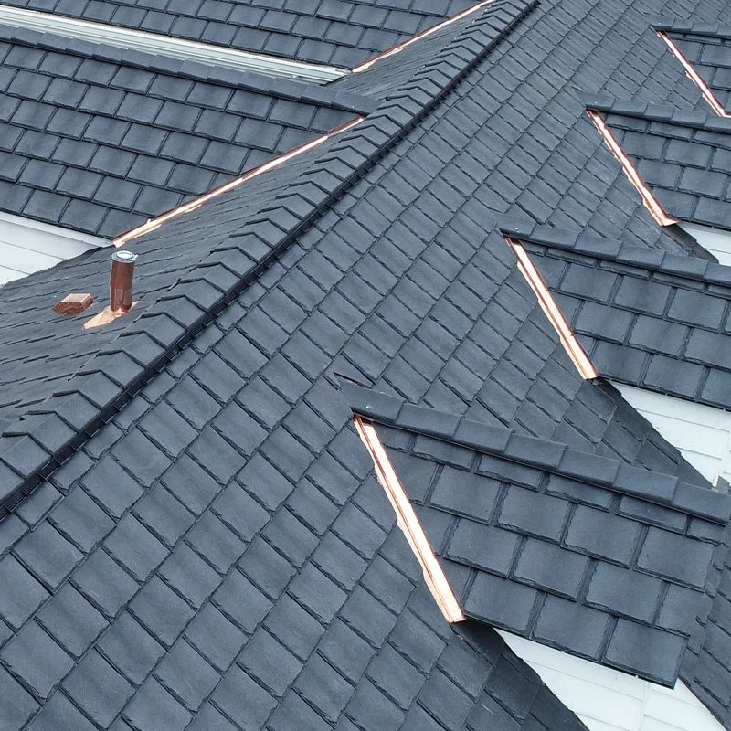 A close-up view of a roof with dark gray asphalt shingles and copper flashing along the edges and around the chimney pipe.