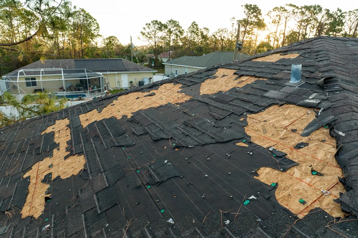 A damaged roof with missing and loose shingles, exposed plywood sheathing, and some old shingles still in place, during sunset in a suburban neighborhood.