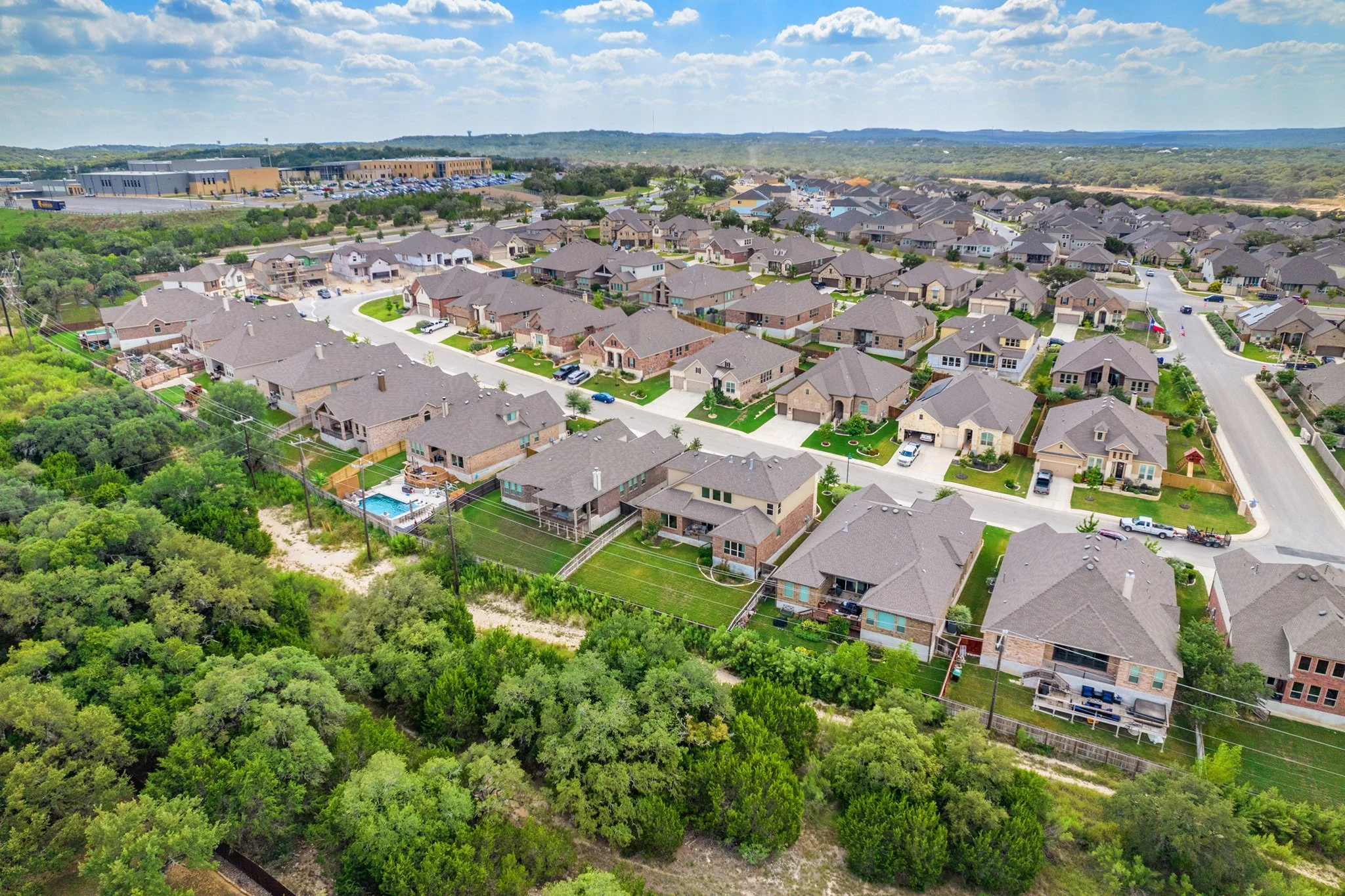 Aerial view of a suburban neighborhood with single-family homes, streets, and green lawns, with wooded area in the foreground and commercial buildings in the distance under partly cloudy skies.