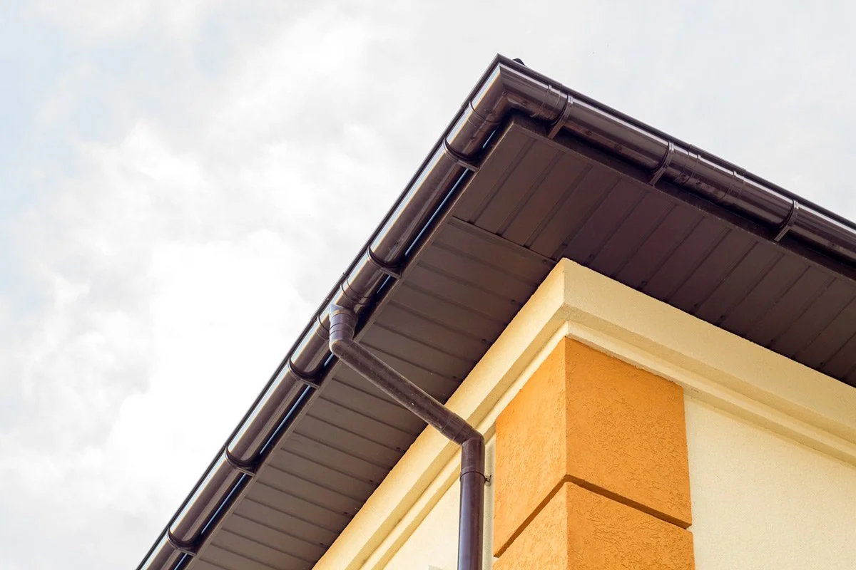 Corner of a house with dark brown rain gutter and downspout, beige and orange exterior walls, and cloudy sky in the background.