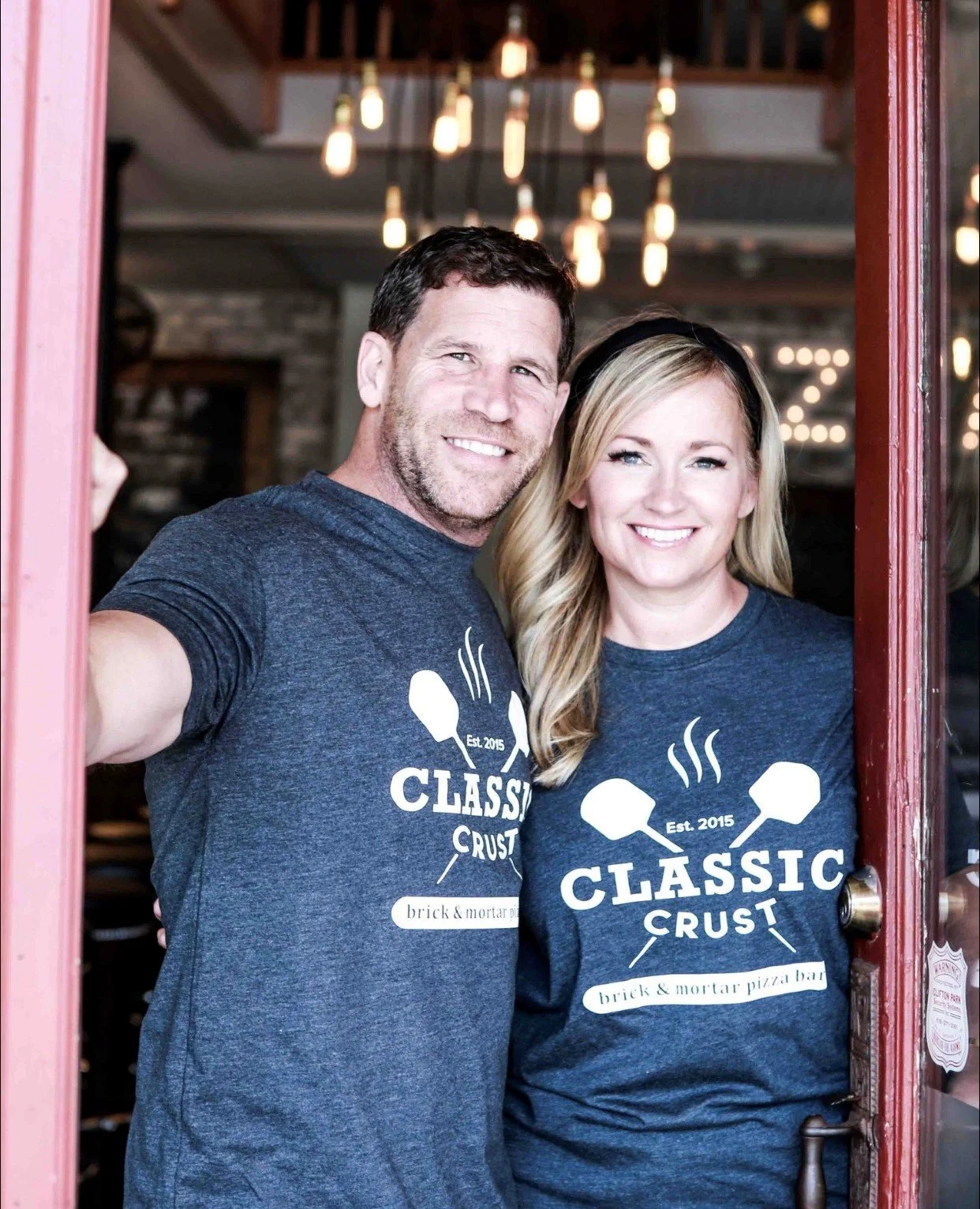 Two smiling people wearing matching dark blue t-shirts with a logo that says "Classic Crust" and "brick & mortar pizza bar" are standing at a doorway, taking a selfie.
