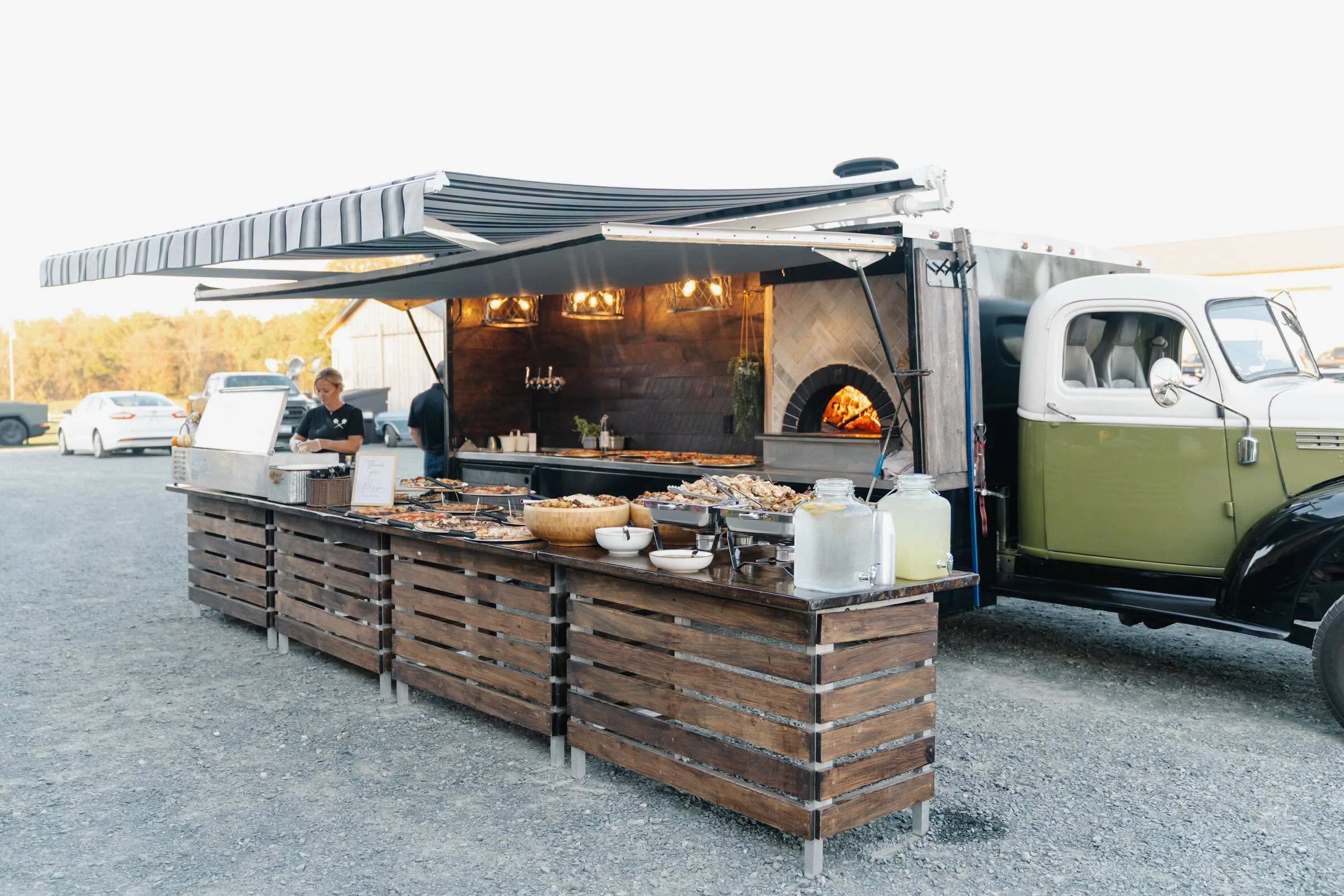 Food truck with outdoor serving area and pizza on display, parked on gravel with multiple cars in the background.
