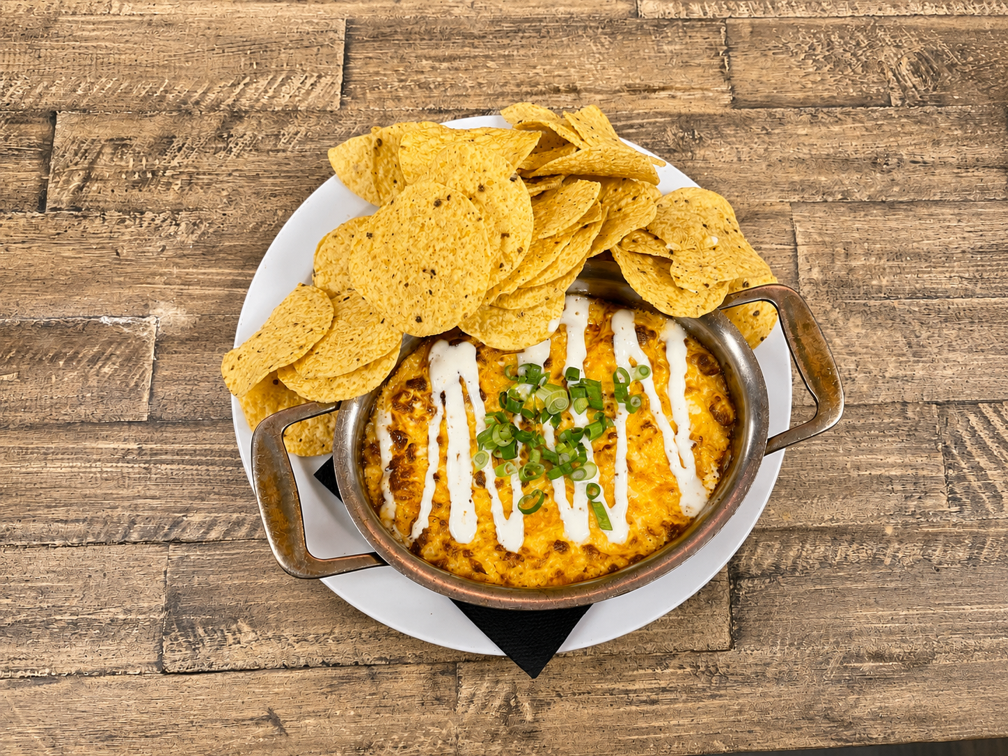 Baked cheesy dip garnished with green onions, served in a small metal dish with sliced tortilla chips on a white plate
