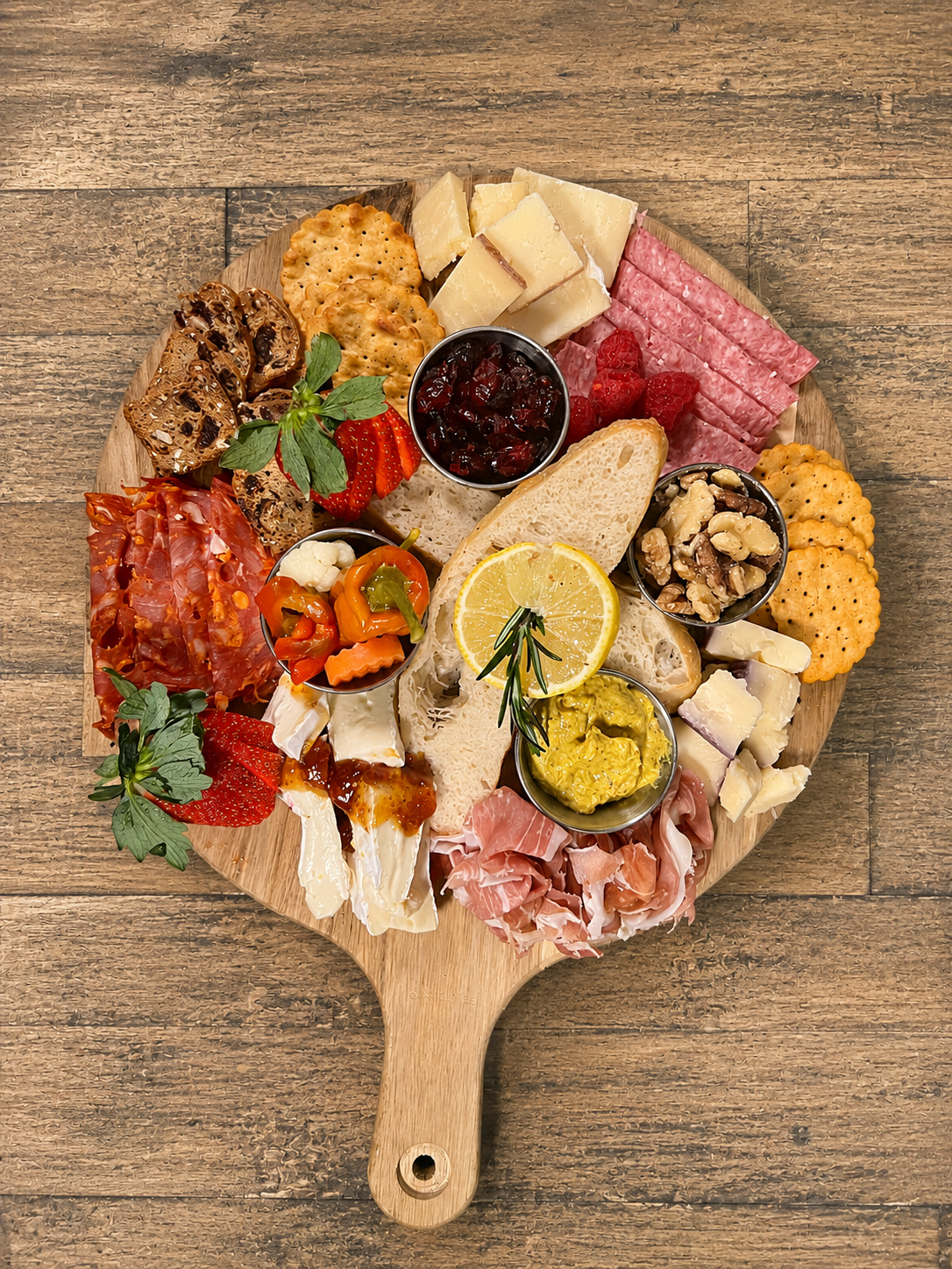 Cheese, crackers, cured meats, fruit preserves, sliced fruits, nuts, and herbs on a wooden serving board.