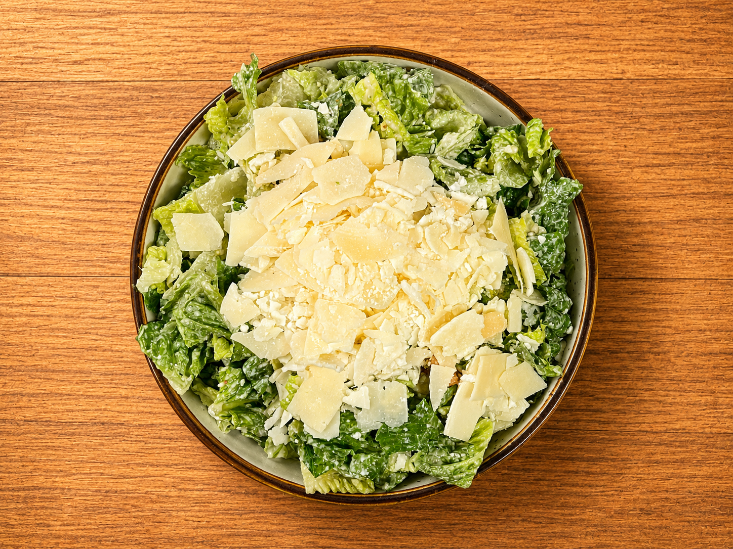 Bowl of Caesar salad topped with shredded cheese on a wooden surface.