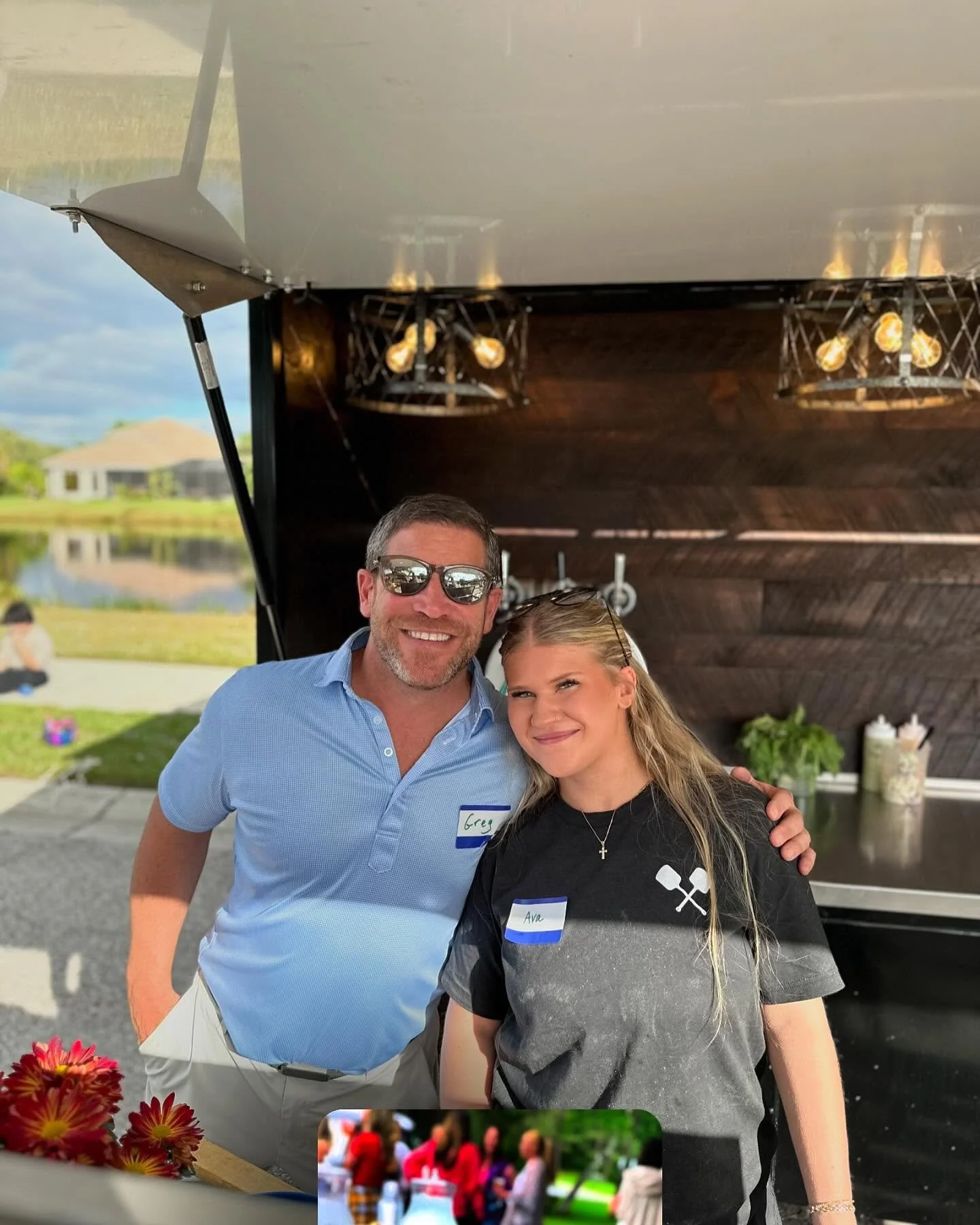 A man and a woman standing side by side, smiling at the camera in an outdoor setting, with a black food truck in the background. The man is wearing sunglasses and a light blue shirt, while the woman has long blonde hair and is wearing a black shirt with a graphic of two spatulas, and a cross necklace. They are both wearing name tags, with the man's tag reading 'Greg' and the woman's reading 'Ava'.