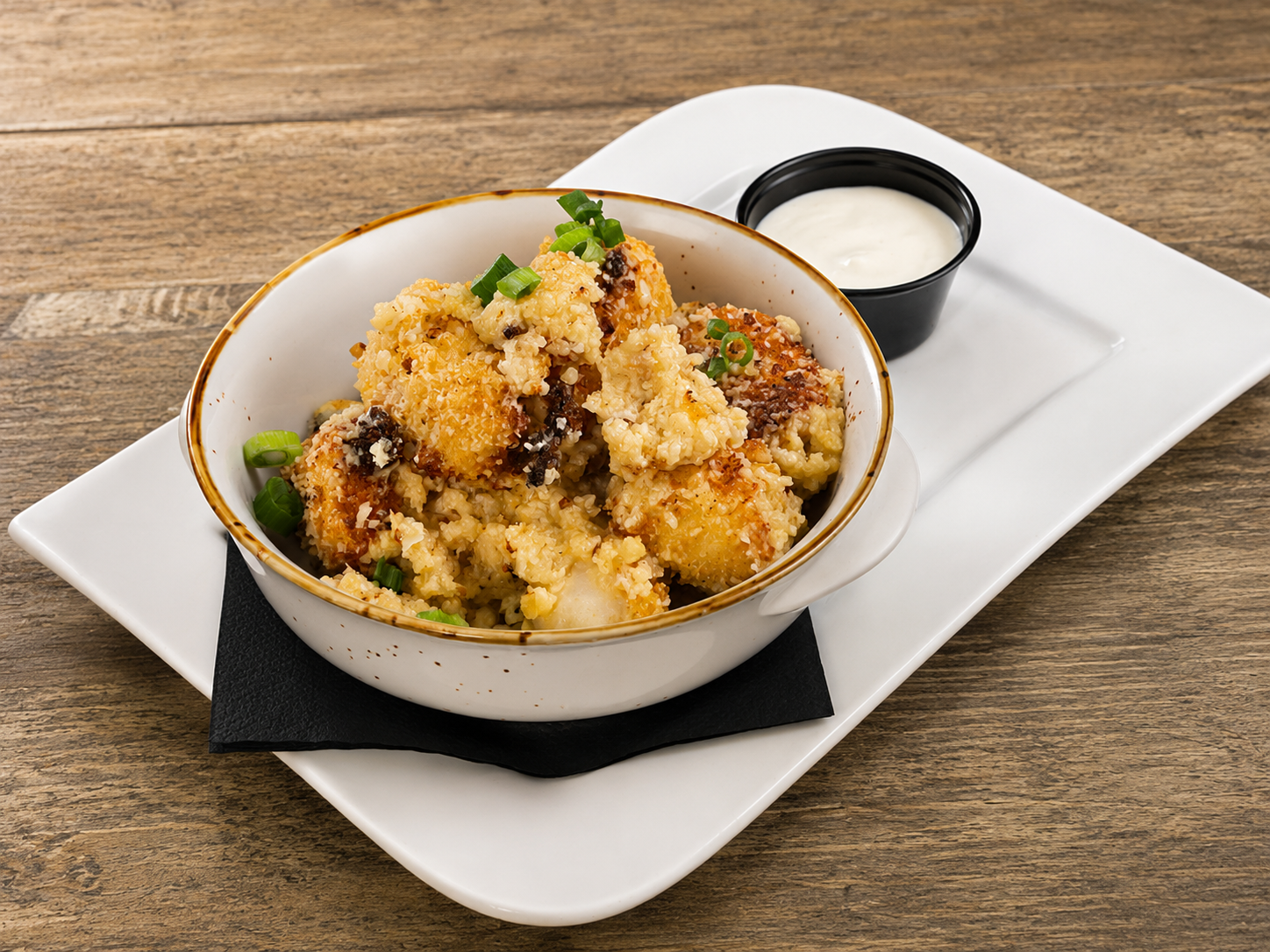 Baked cauliflower bites garnished with chopped green onions served with a side of creamy dipping sauce on a white rectangular plate.