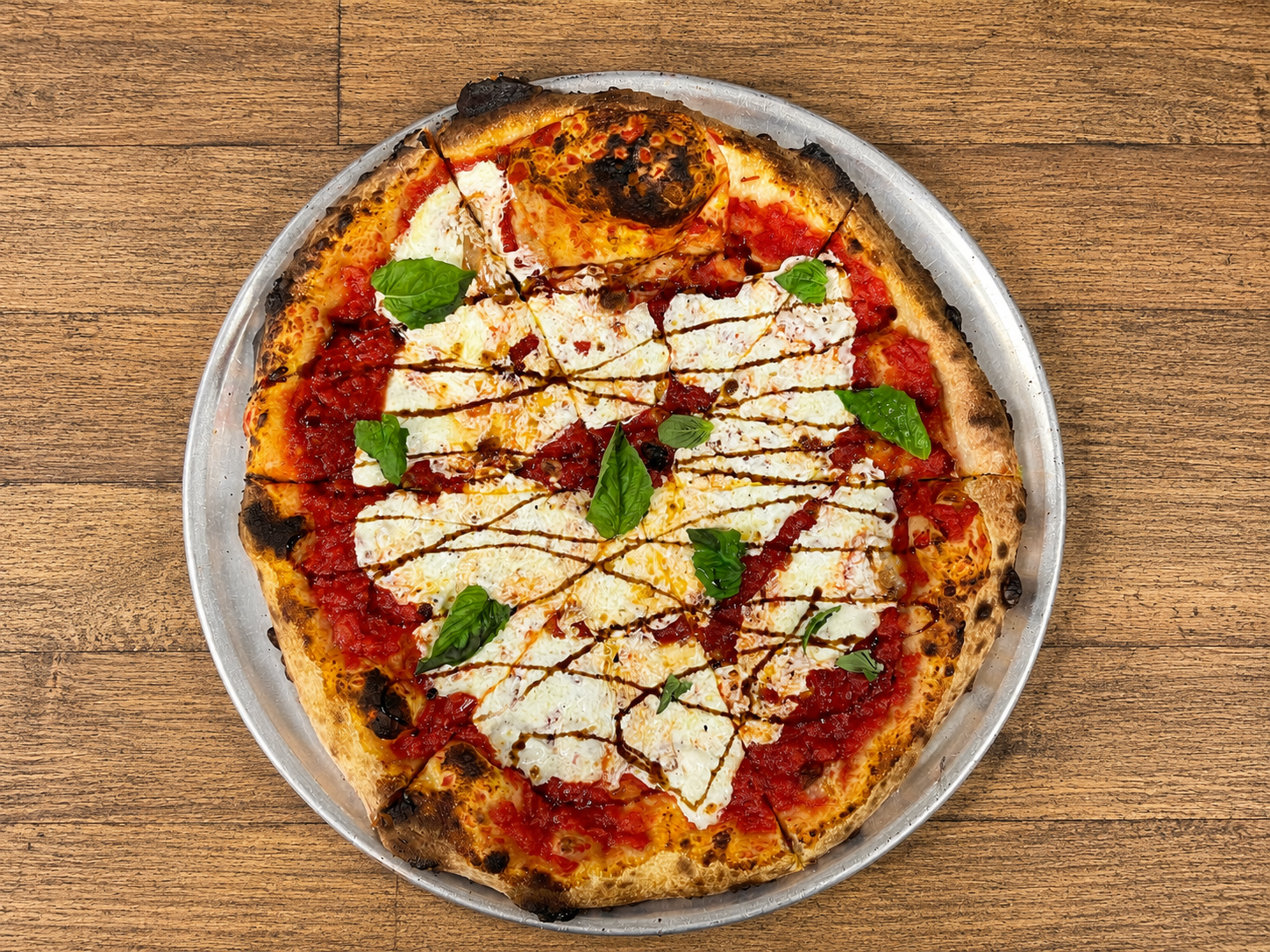 A pizza topped with mozzarella cheese, tomato sauce, fresh basil leaves, and a drizzle of balsamic glaze on a silver tray, placed on a wooden table.