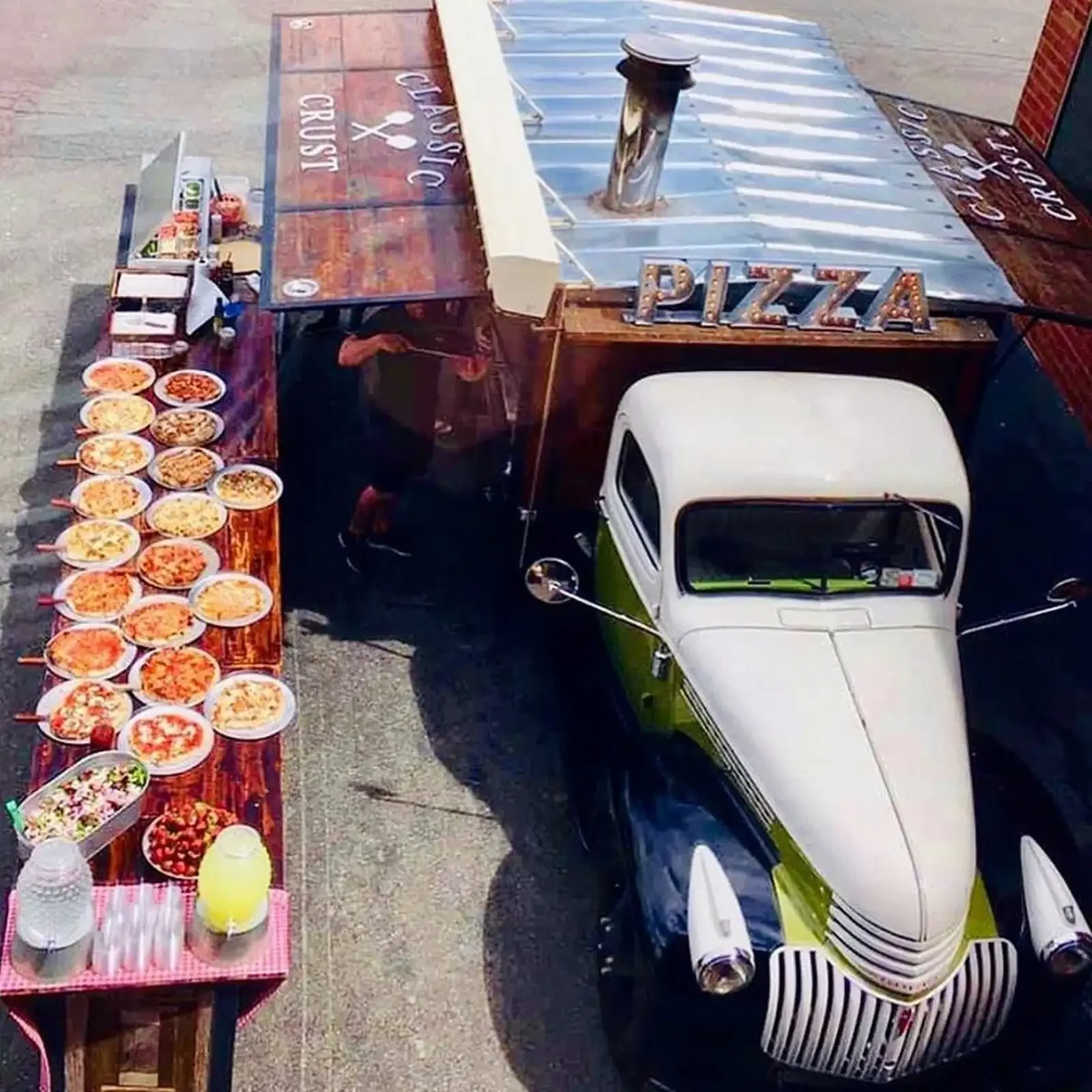 A vintage green and white plane parked next to a pizza stand with multiple bowls of pizza toppings on a wooden table.